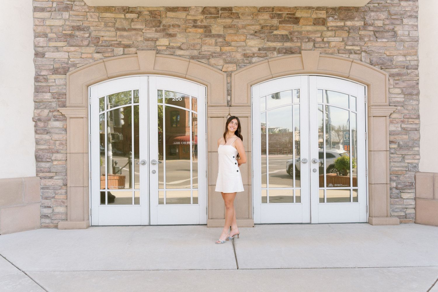 A woman in a white dress poses against a stone wall exterior with arched windows.