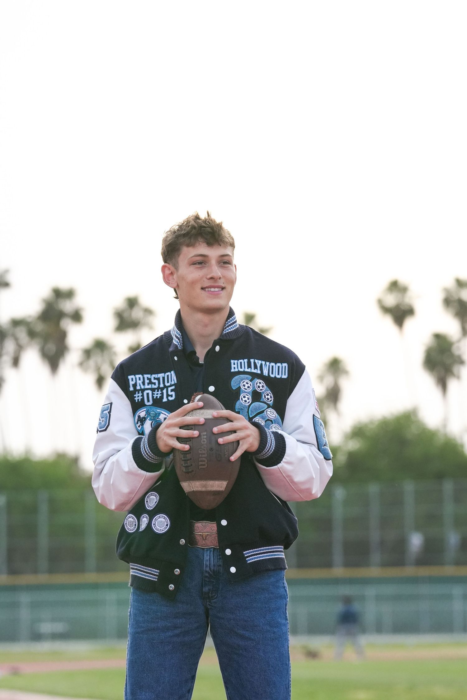 Student athlete poses with football in varsity jacket on sports field.