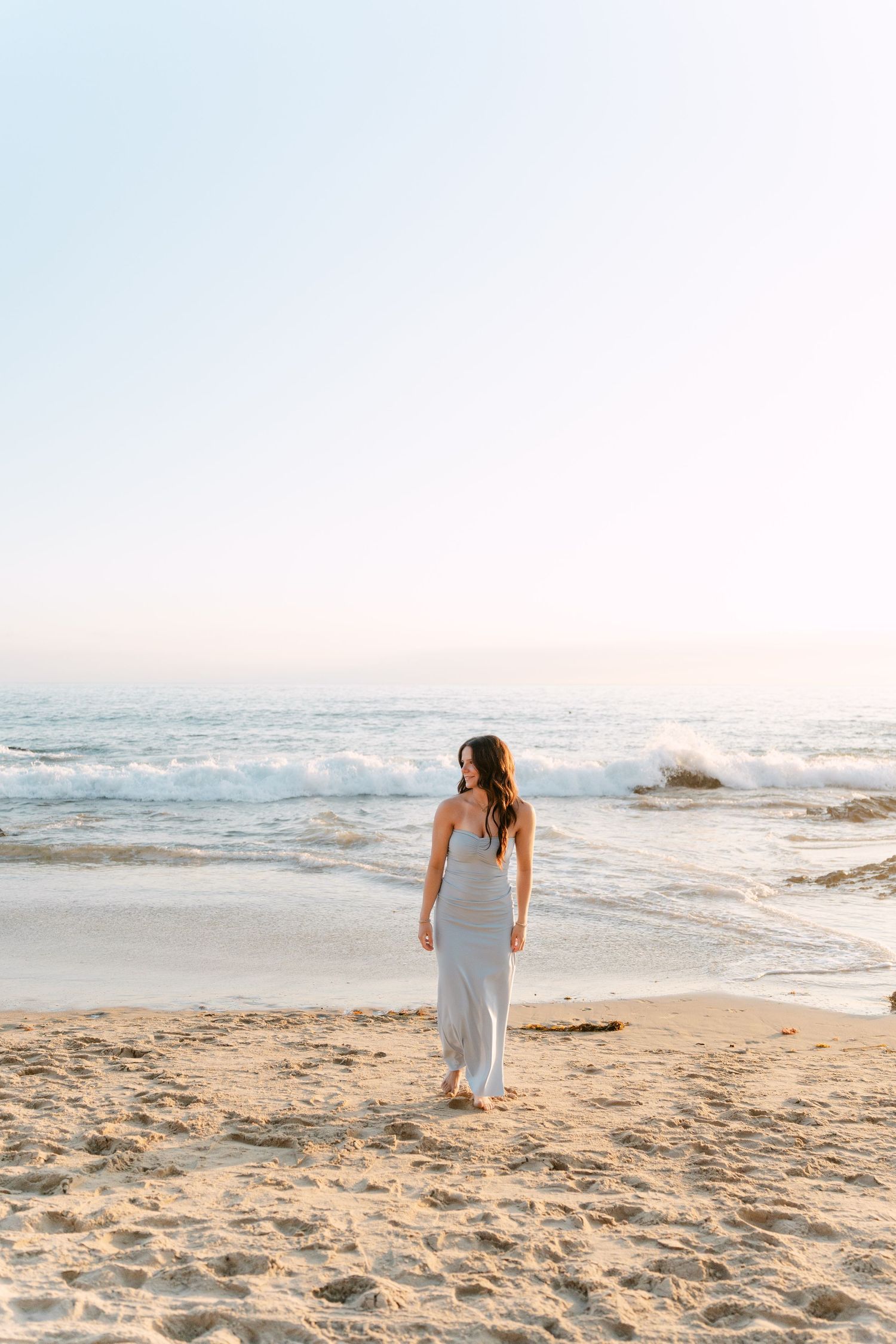 Woman in gray maxi dress standing barefoot on sandy beach, gazing at ocean waves during golden hour sunset.