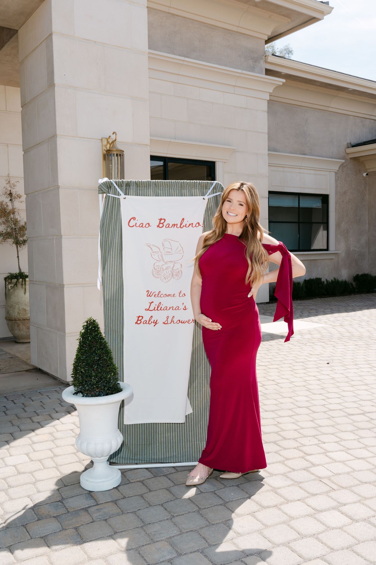 Pregnant woman in red gown poses at Liliana's Baby Shower welcome sign reading Ciao Bambino outdoors.