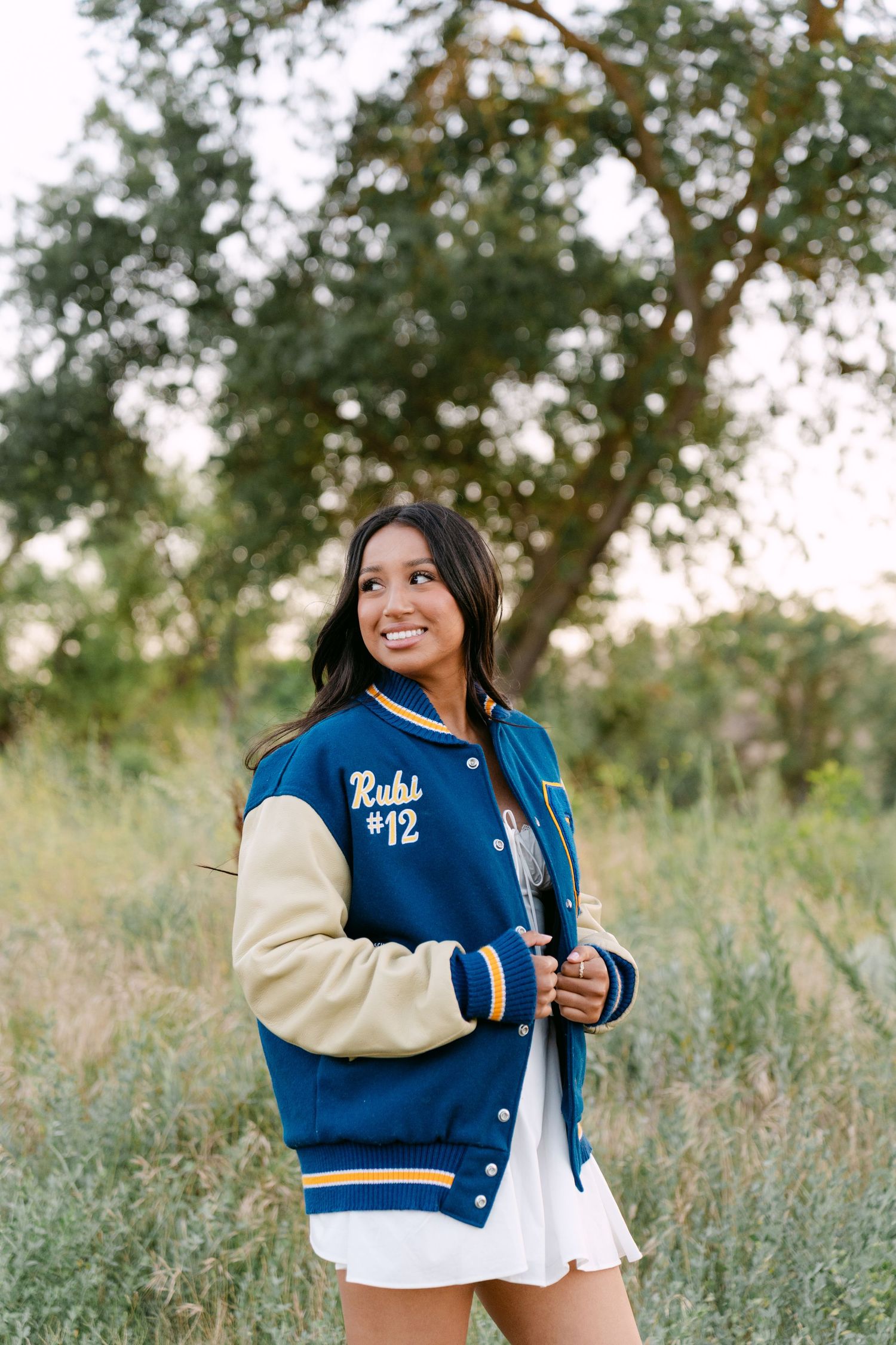 Smiling teen in blue varsity jacket numbered 12 poses outdoors in green field for senior portrait photo.