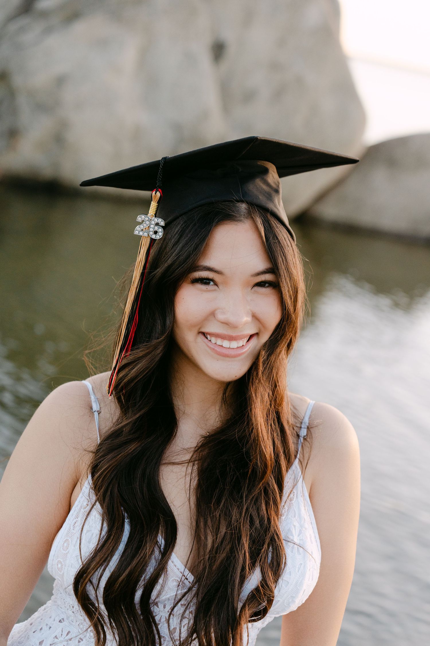 Smiling young woman wearing a black graduation cap and white dress, posing outdoors near water and rocks.