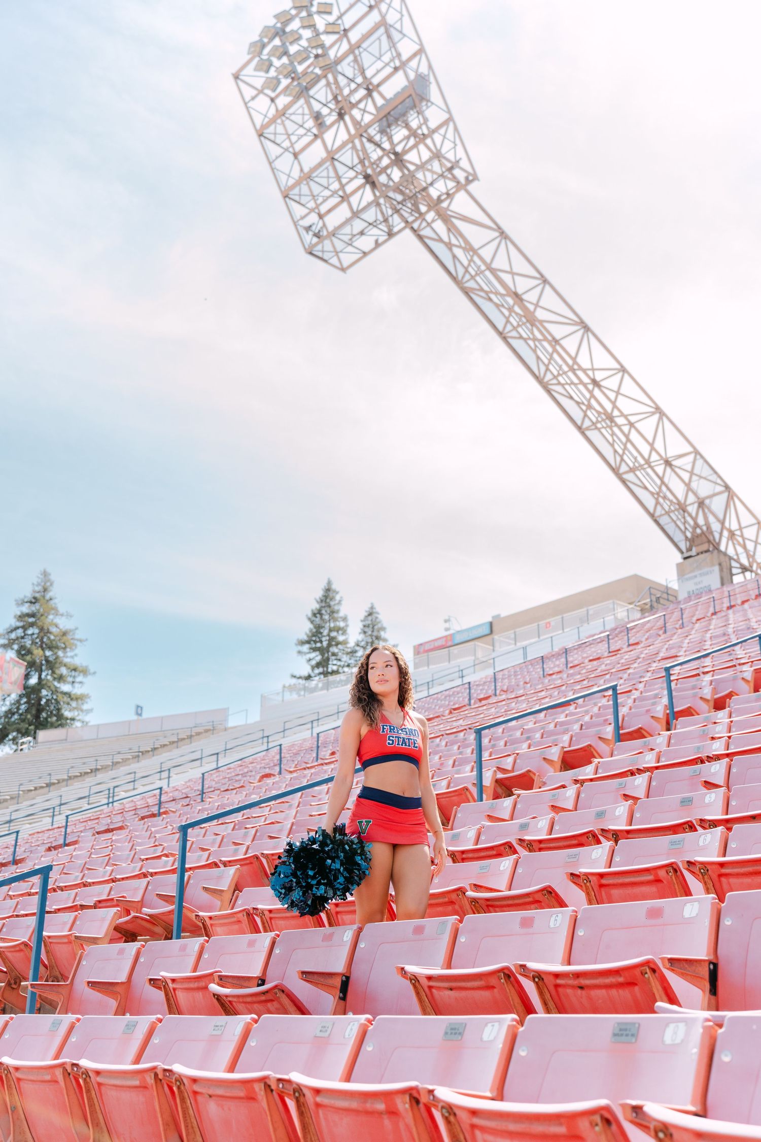 Cheerleader in red uniform stands in empty red stadium bleachers holding pom-poms beneath stadium lights.