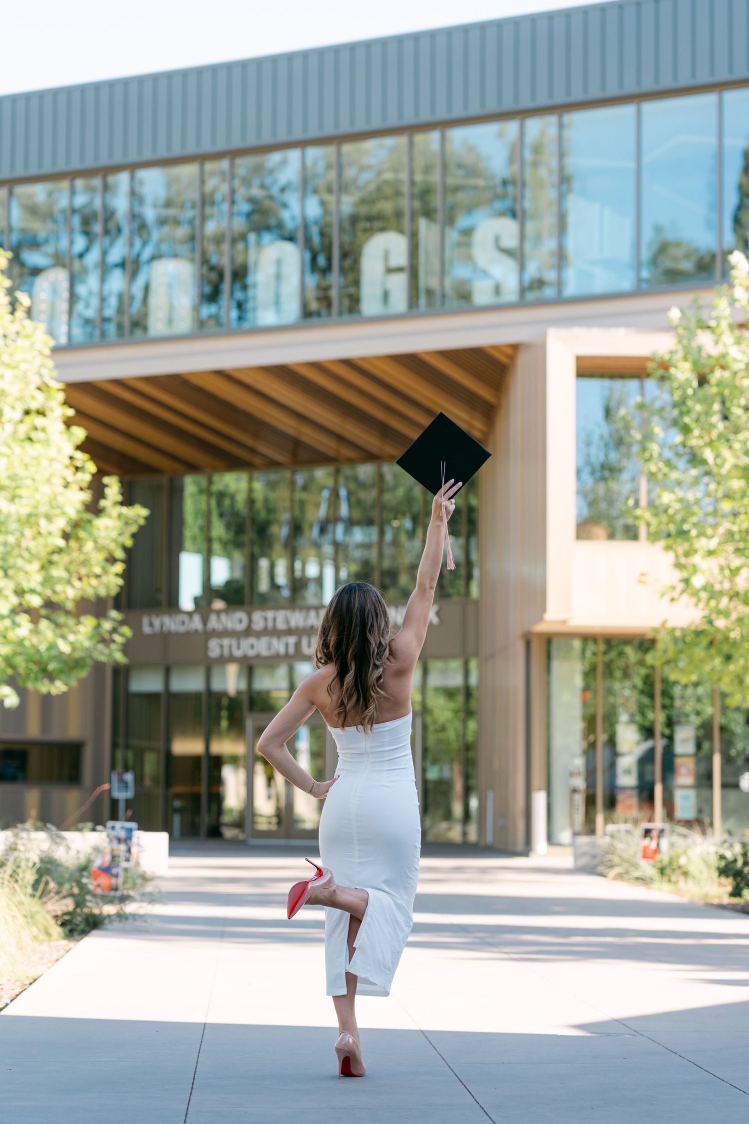Graduate in white dress tosses cap outside Lynda and Stewart Student Union building on sunny day.
