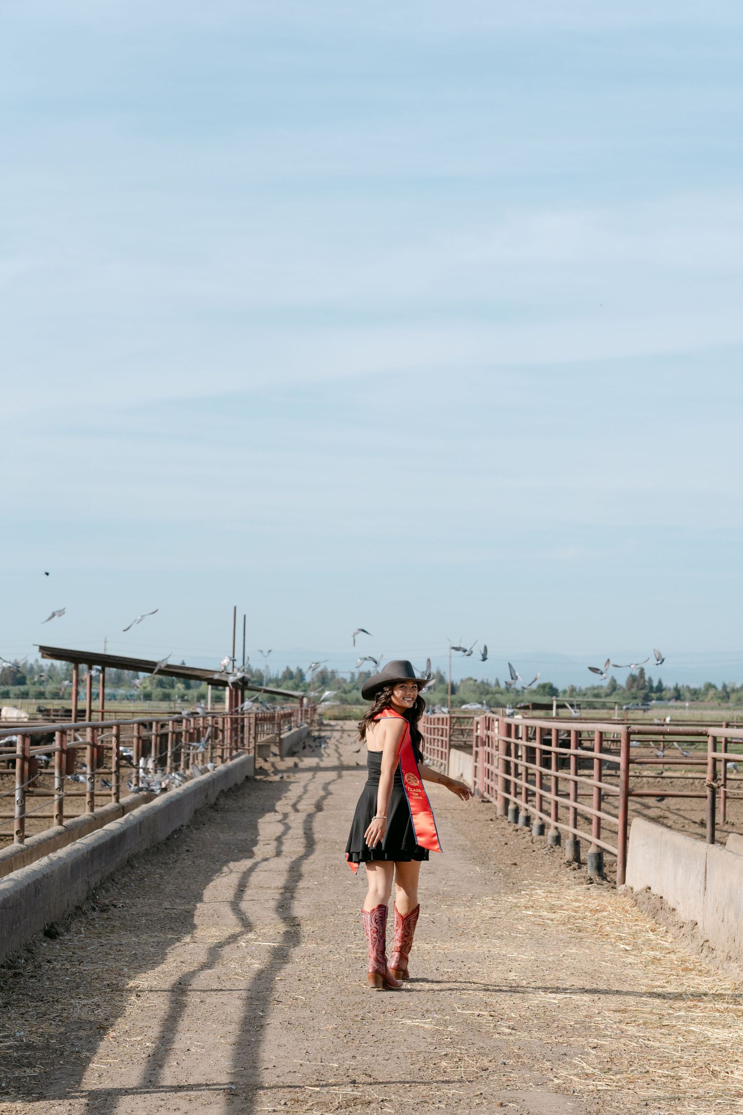 Woman in black dress, red shawl, cowboy hat and boots poses on a ranch pathway with cattle pens on a sunny day.