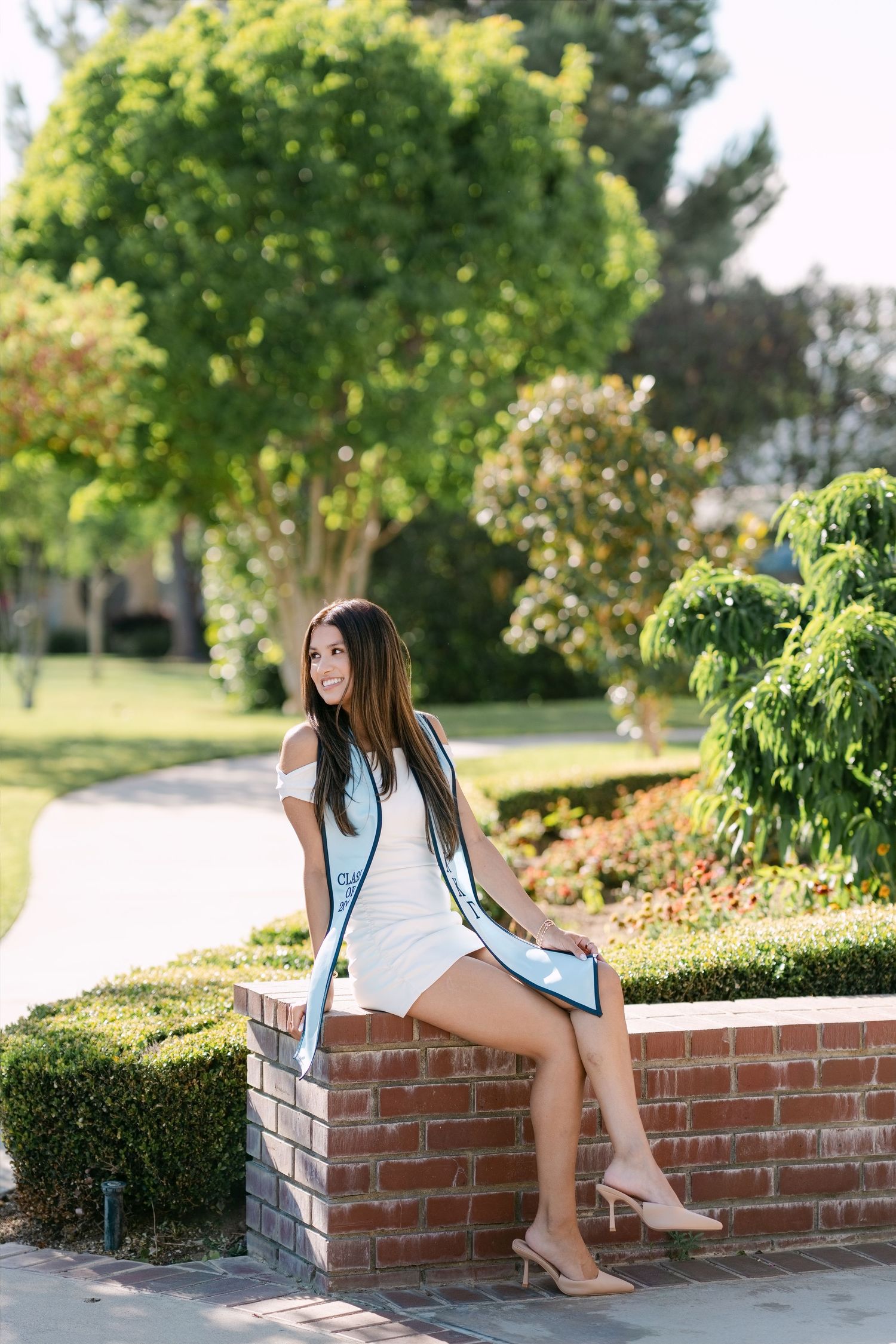 Young woman in white dress with light blue graduation sash sits on brick wall in sunny garden, smiling.