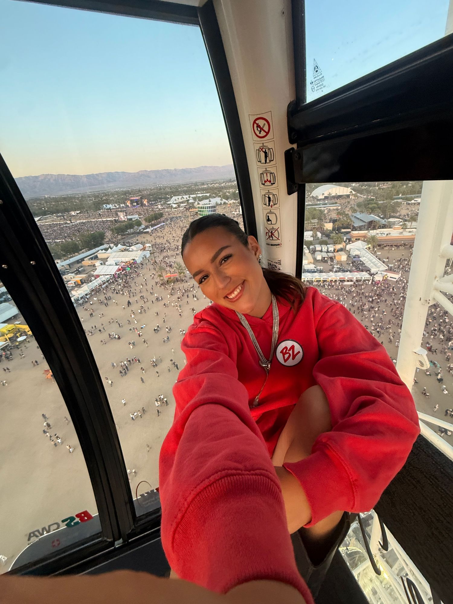 Young woman in red hoodie smiling in a gondola ride high above a crowded outdoor festival with mountain views.