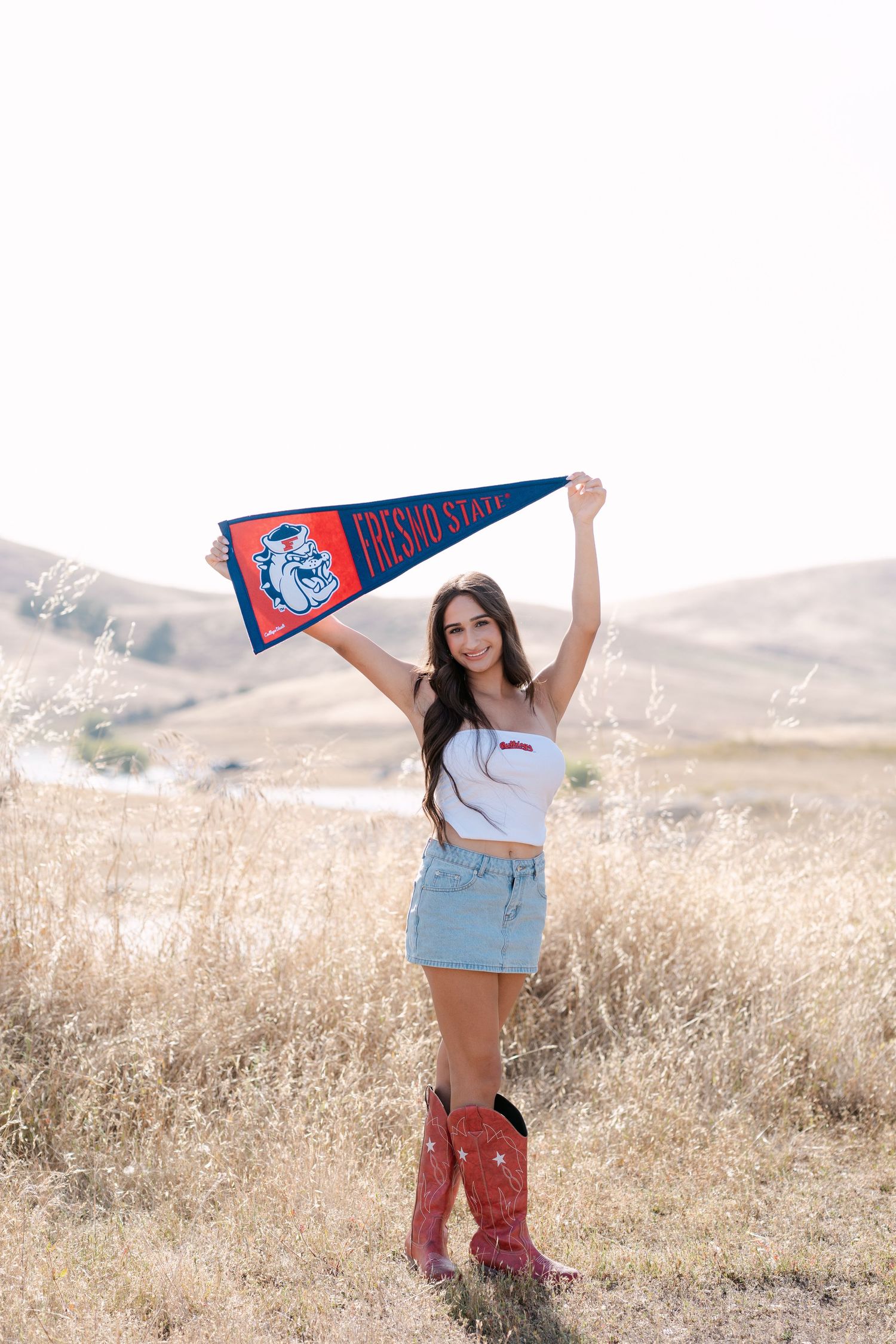 Young woman holding a Fresno State pennant in a field, wearing a denim skirt and red cowboy boots.