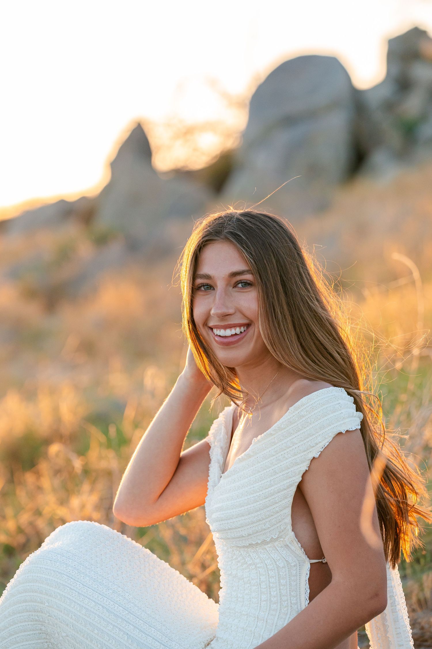 Smiling woman in white dress sitting outdoors at golden hour with rocky landscape in background.
