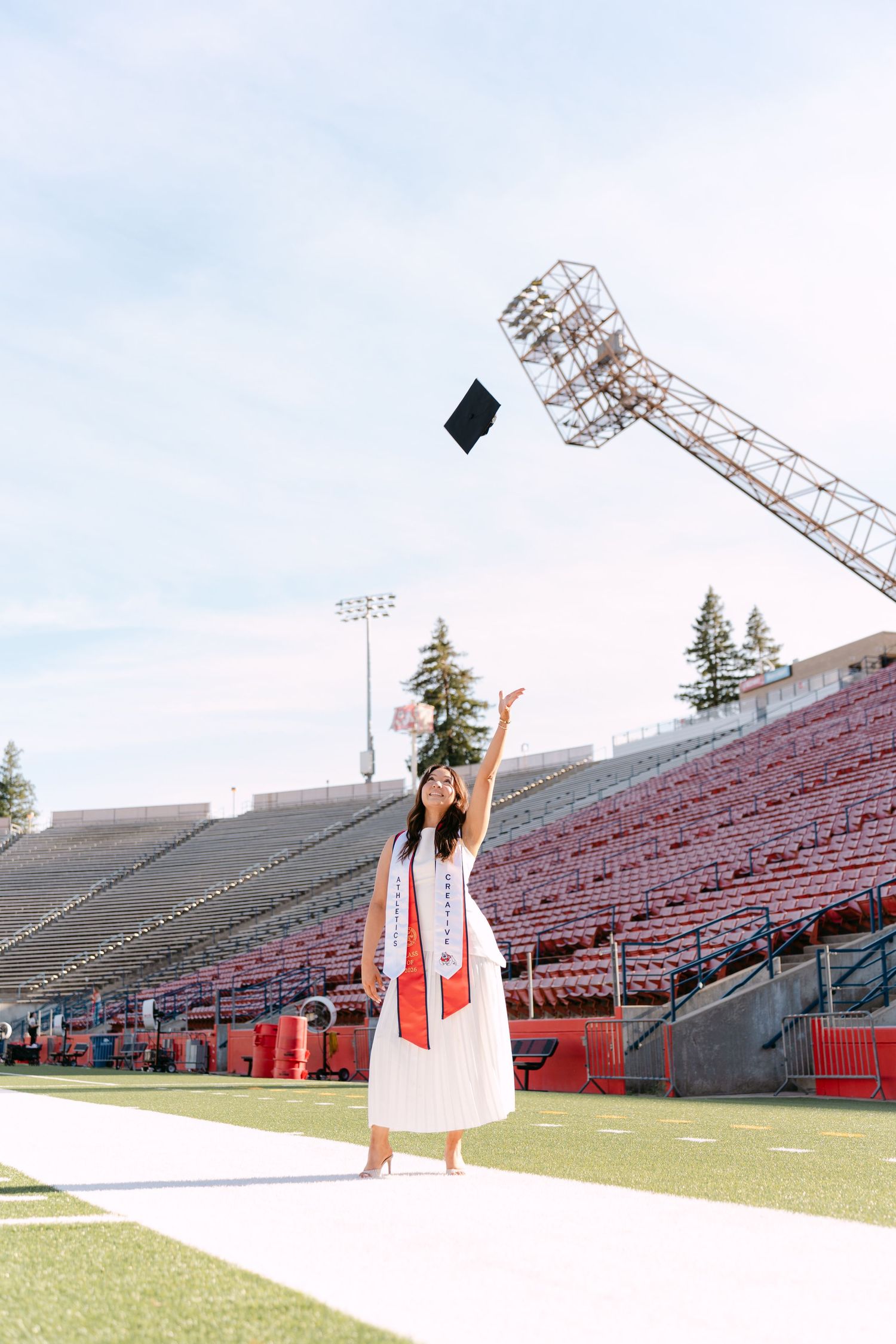 Graduate in white gown tossing cap in stadium stands, celebrating graduation day outdoors.