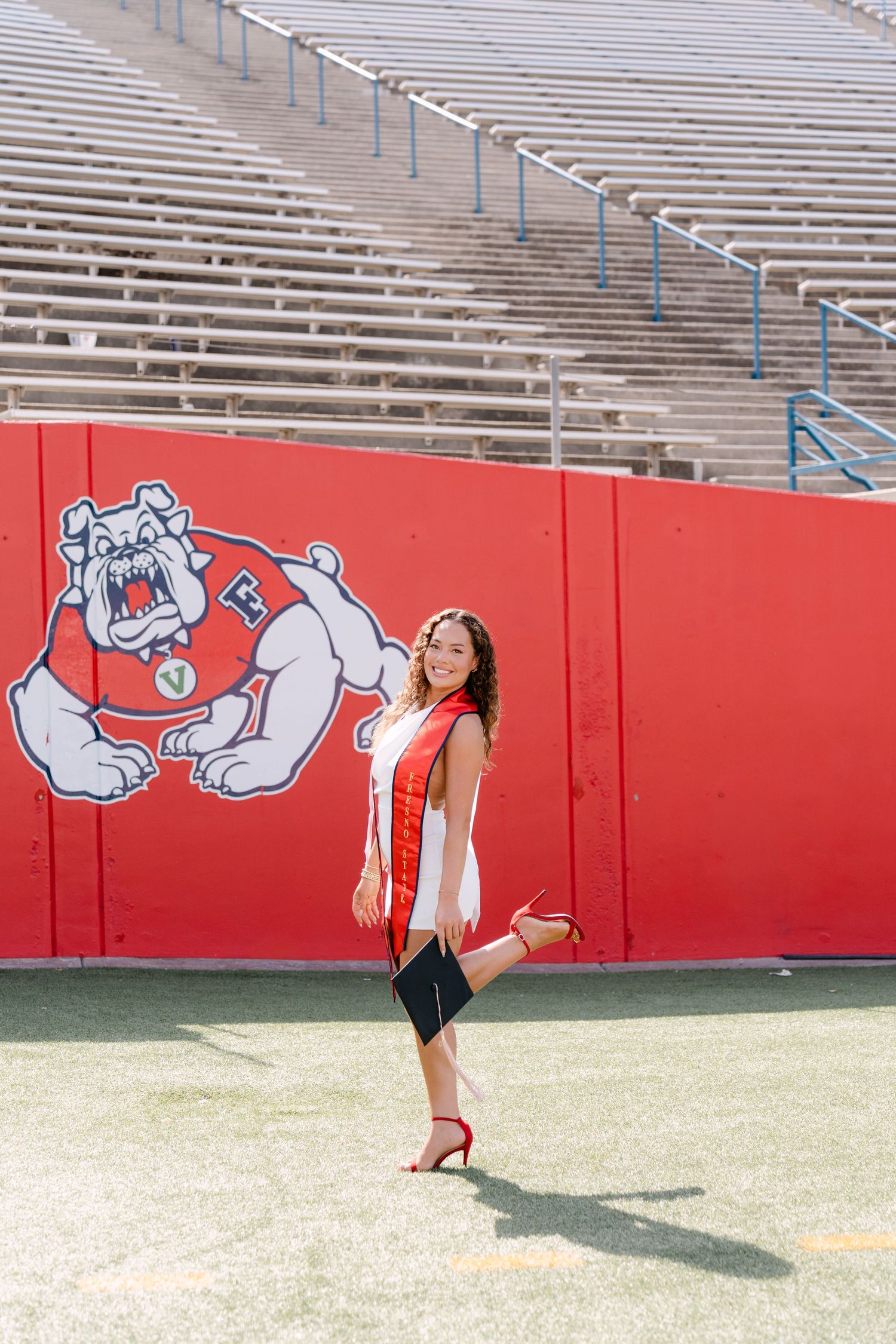 Graduate poses in red sash at Fresno State Bulldog stadium, holding diploma, wearing red heels on game field.