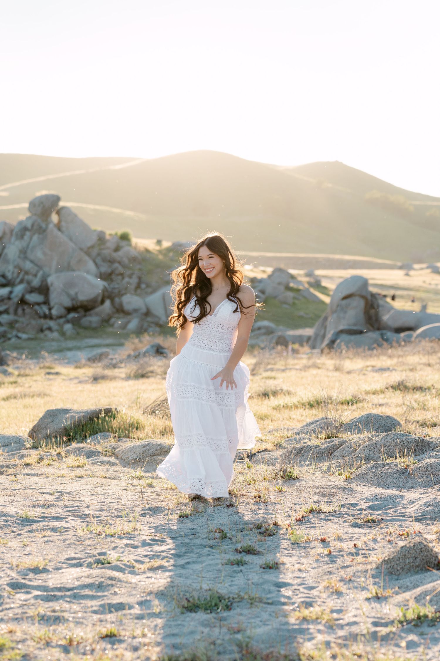 Young woman in white lace dress poses gracefully in sunlit rocky landscape with rolling hills at golden hour.