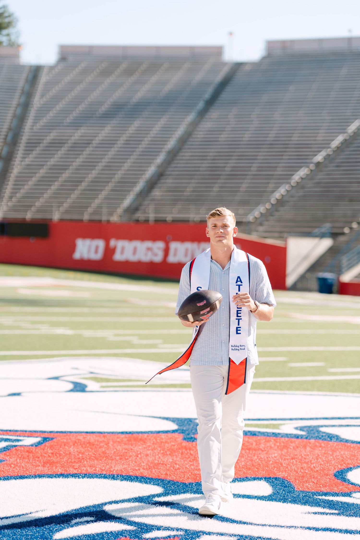 Graduate in white walks on football field holding cap, wearing blue sash, with stadium seating in background.