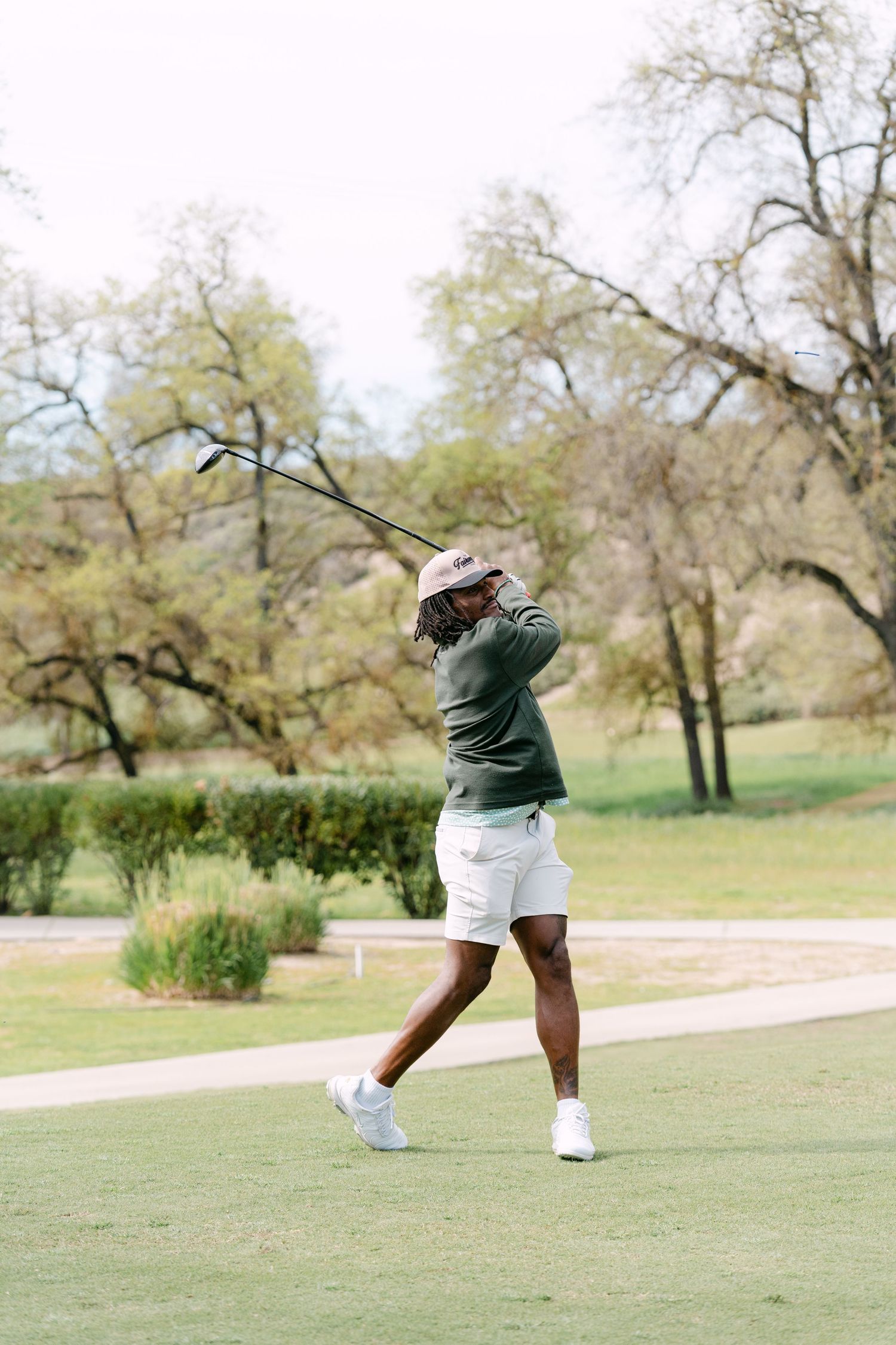 A golfer in white shorts and green sweater completes a swing on a lush golf course surrounded by trees.