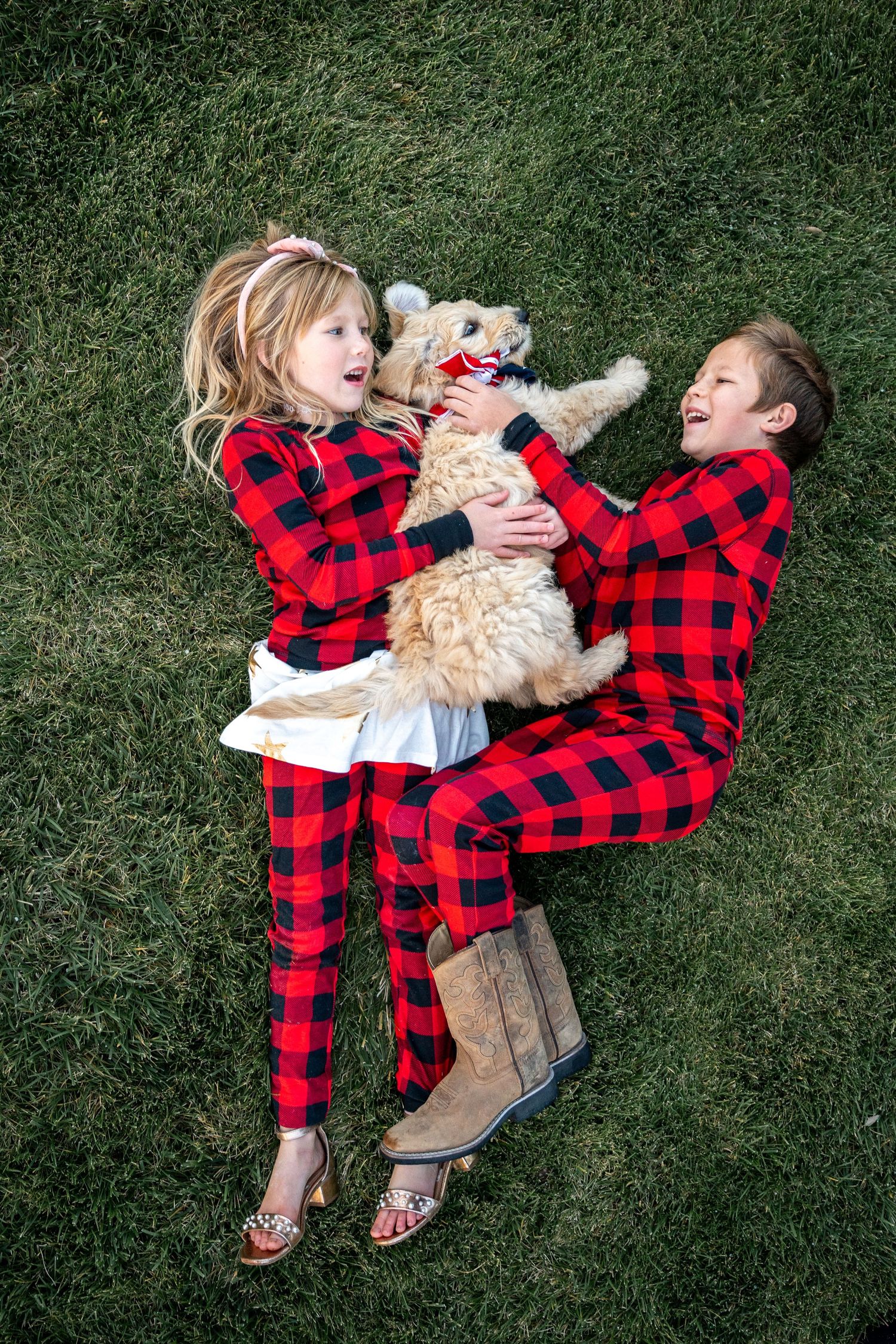 Two people in matching red buffalo plaid outfits playing on grass with a small white dog.