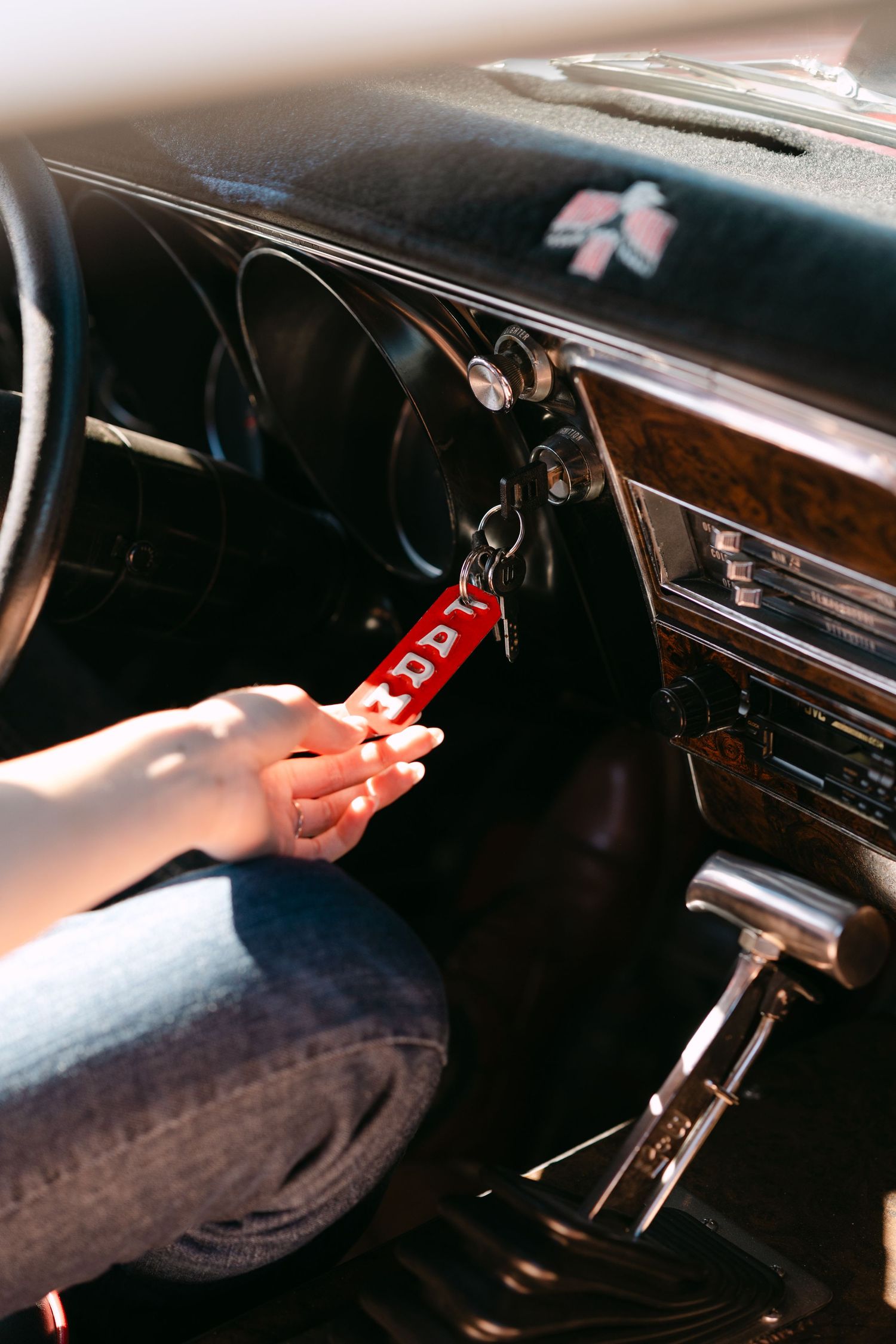 A hand inserts a red keychain key into the ignition of a vintage car with wood grain dashboard.