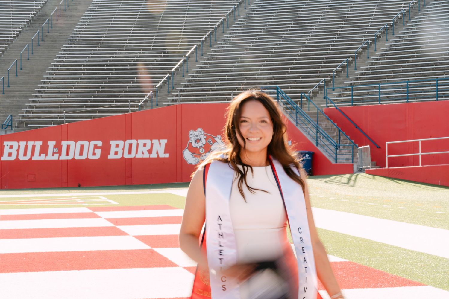 Smiling graduate in white stole poses at Fresno State Bulldog Born stadium on a sunny day.