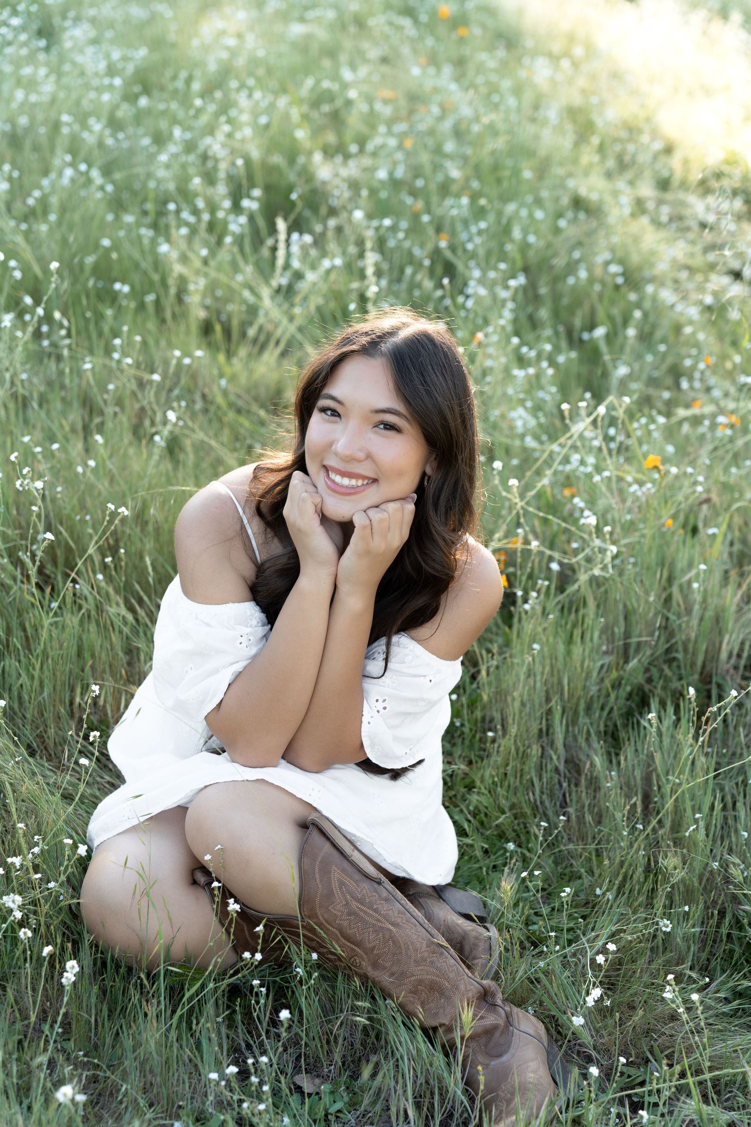 Young woman in white off-shoulder dress and cowboy boots sitting in a wildflower field, smiling cheerfully.