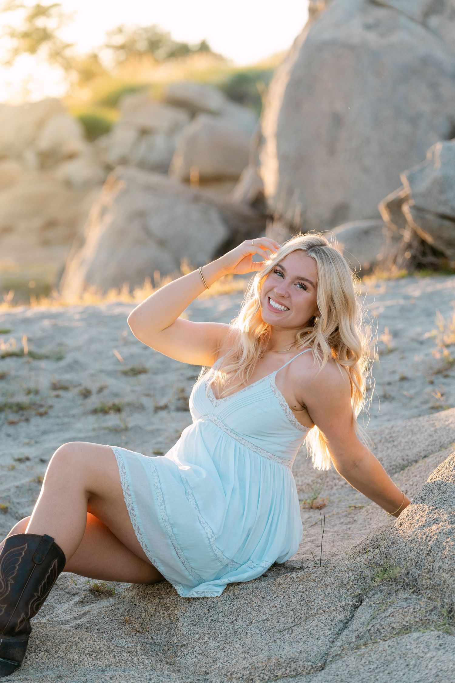 Smiling blonde woman in white sundress and cowboy boots sitting on sandy rocks during golden hour sunset.
