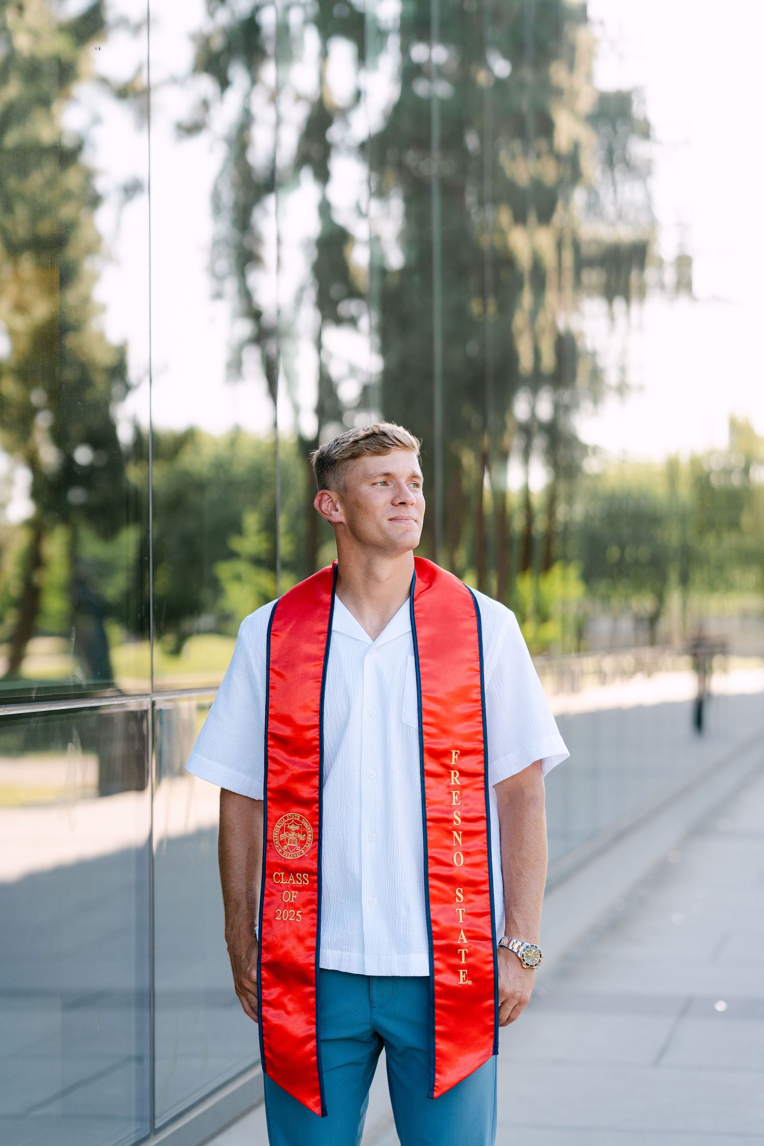 Young man in white shirt and red graduation sash stands confidently outdoors near glass wall with trees reflected behind him.