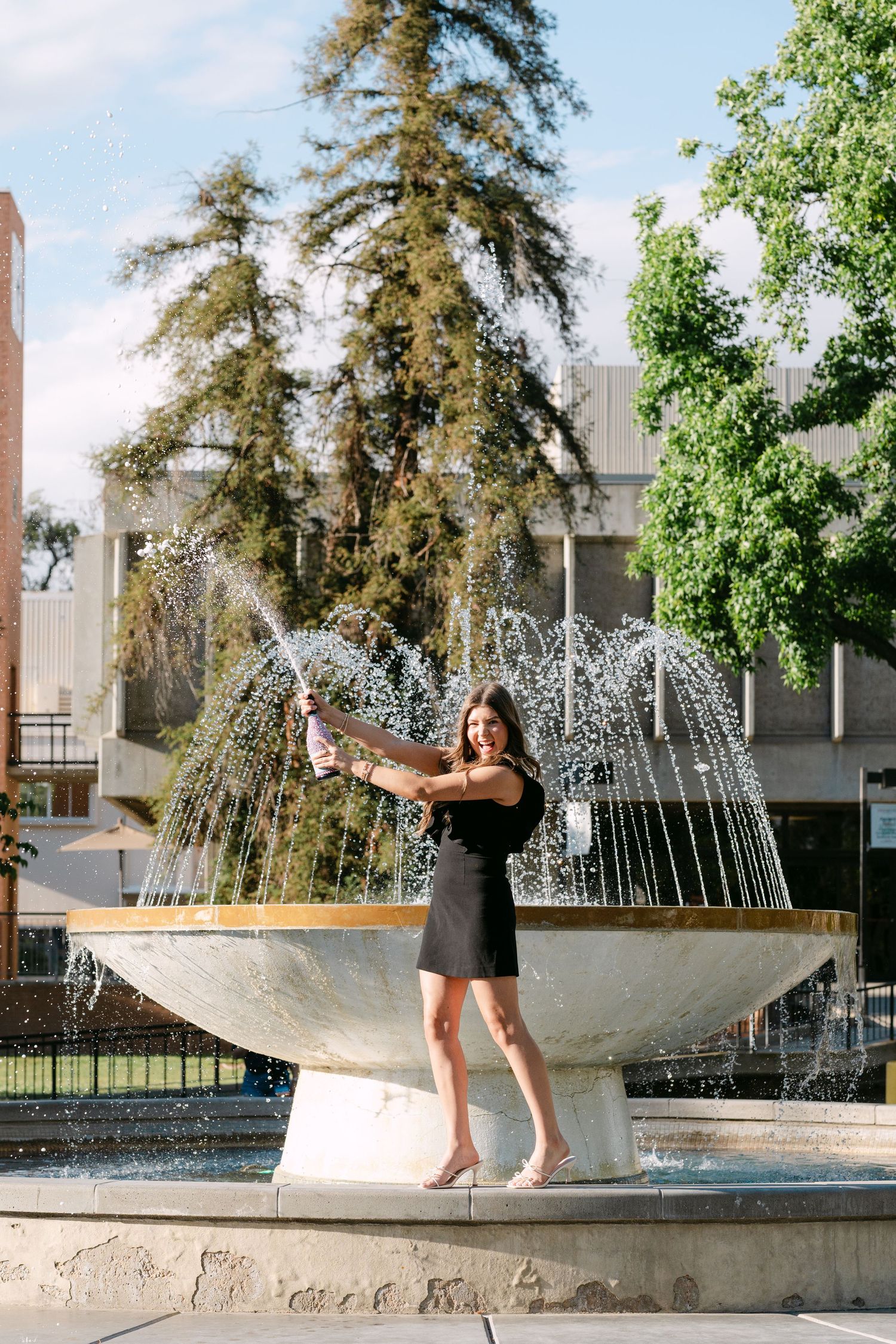 Woman in black dress popping champagne bottle at outdoor fountain with trees in background.