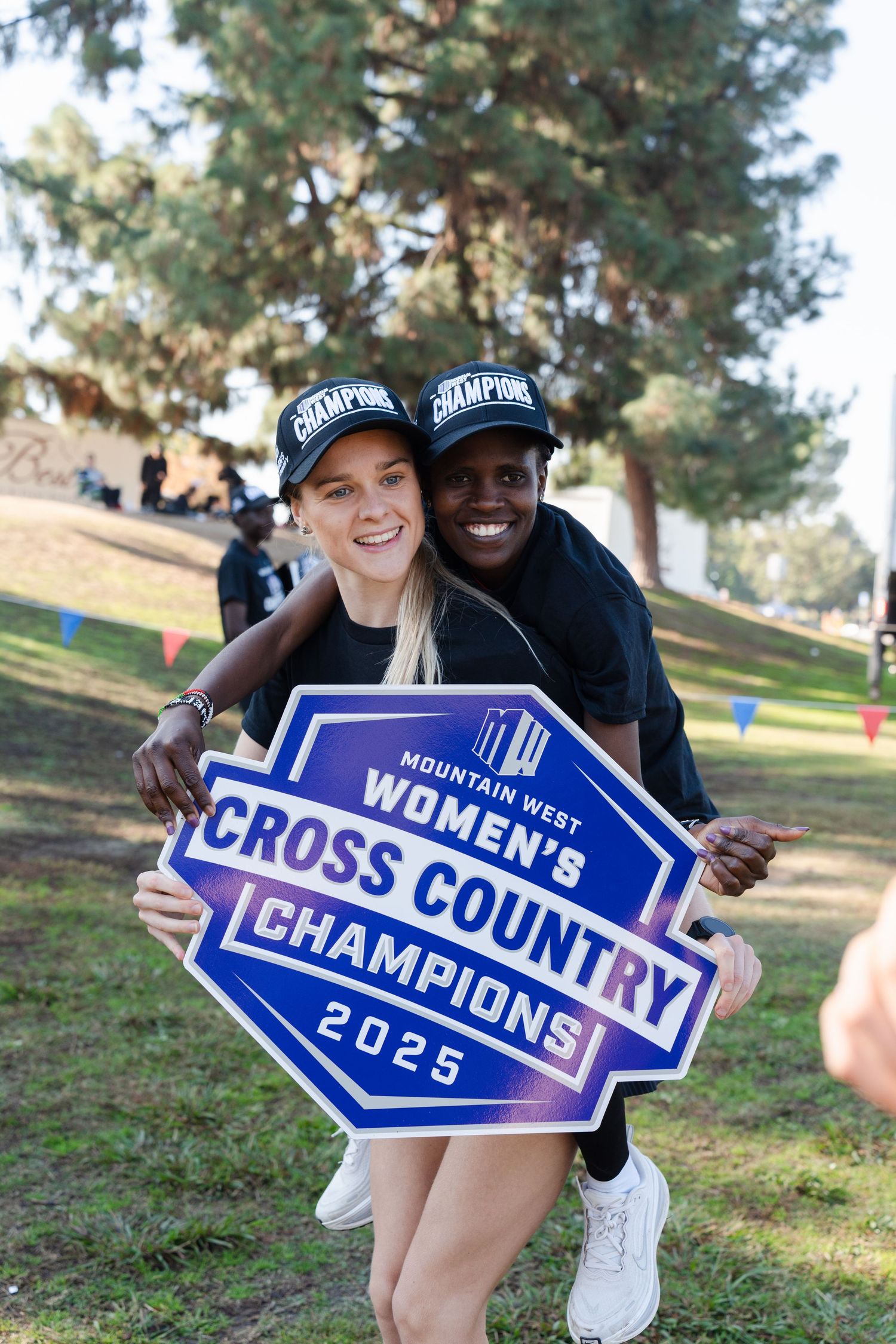 Two athletes pose with blue championship signs on a grassy hill during a sunny day.
