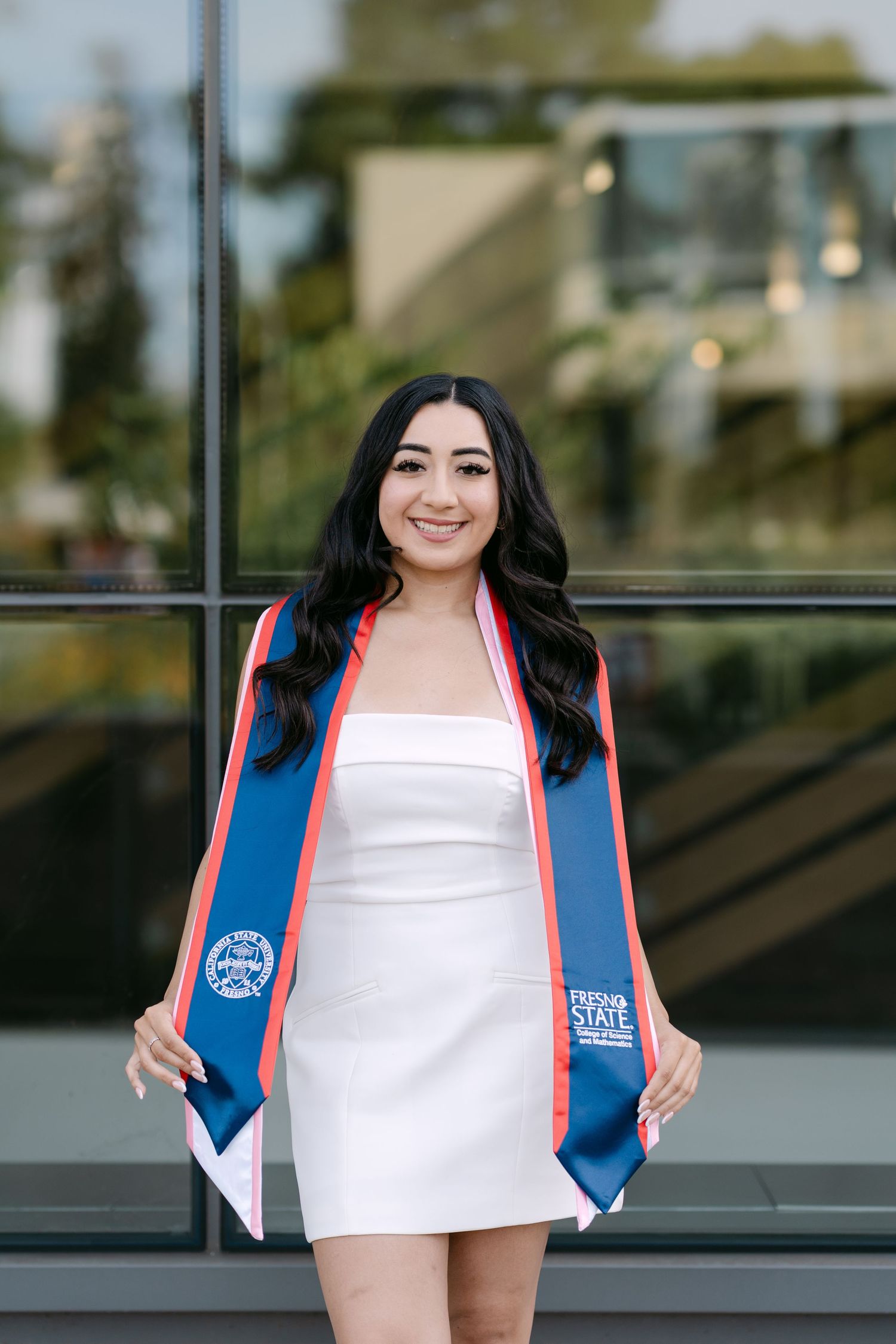 Fresno State graduate in white dress holding blue and red graduation stole, smiling outdoors.