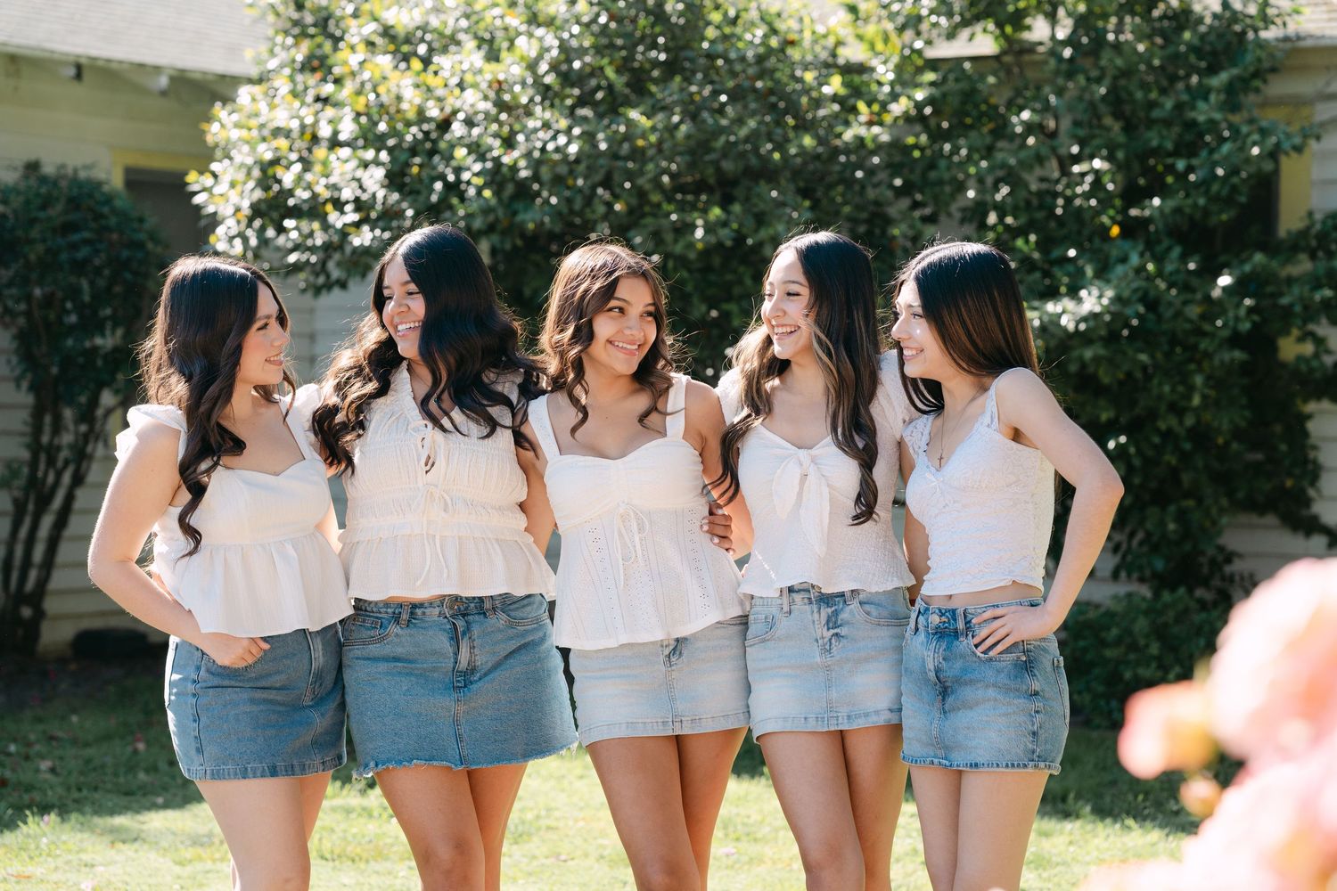Five young women in white tops and denim skirts smiling and laughing together outdoors in a sunny garden.