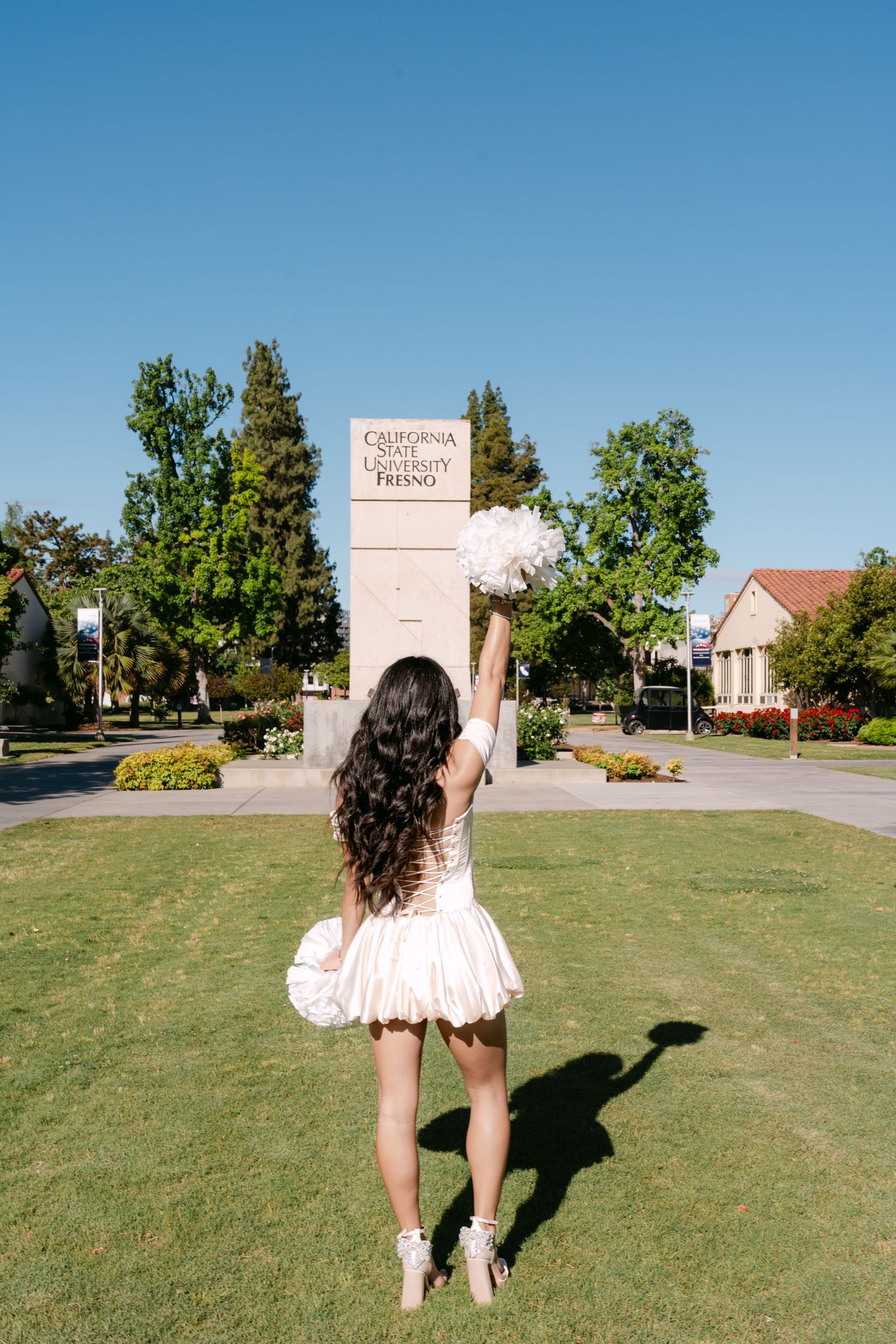 Cheerleader in white uniform raises pom-poms in front of California State University Fresno campus sign on sunny day.