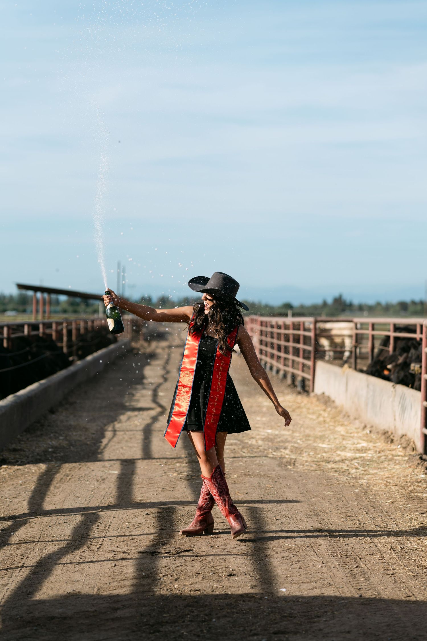 Woman in black dress, cowboy hat, and red boots sprays champagne at a farm with cattle pens under blue sky.