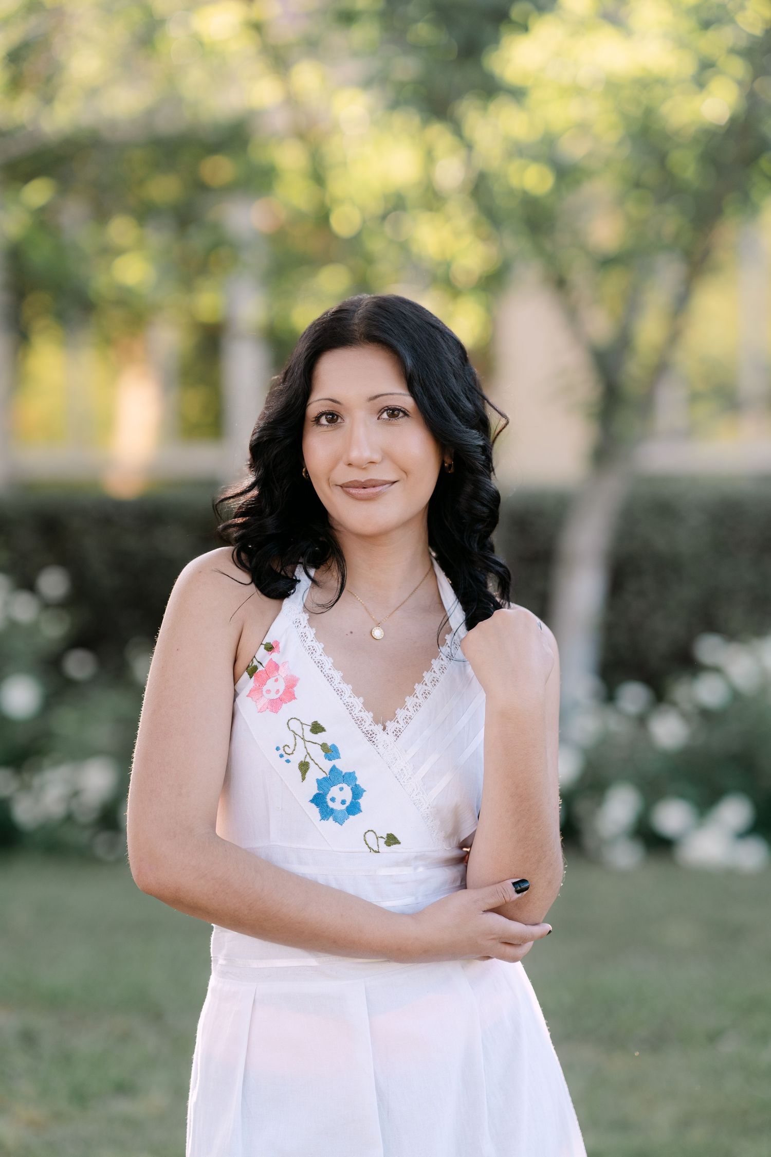 Woman in white floral embroidered dress posing outdoors in a garden with green trees in the background.