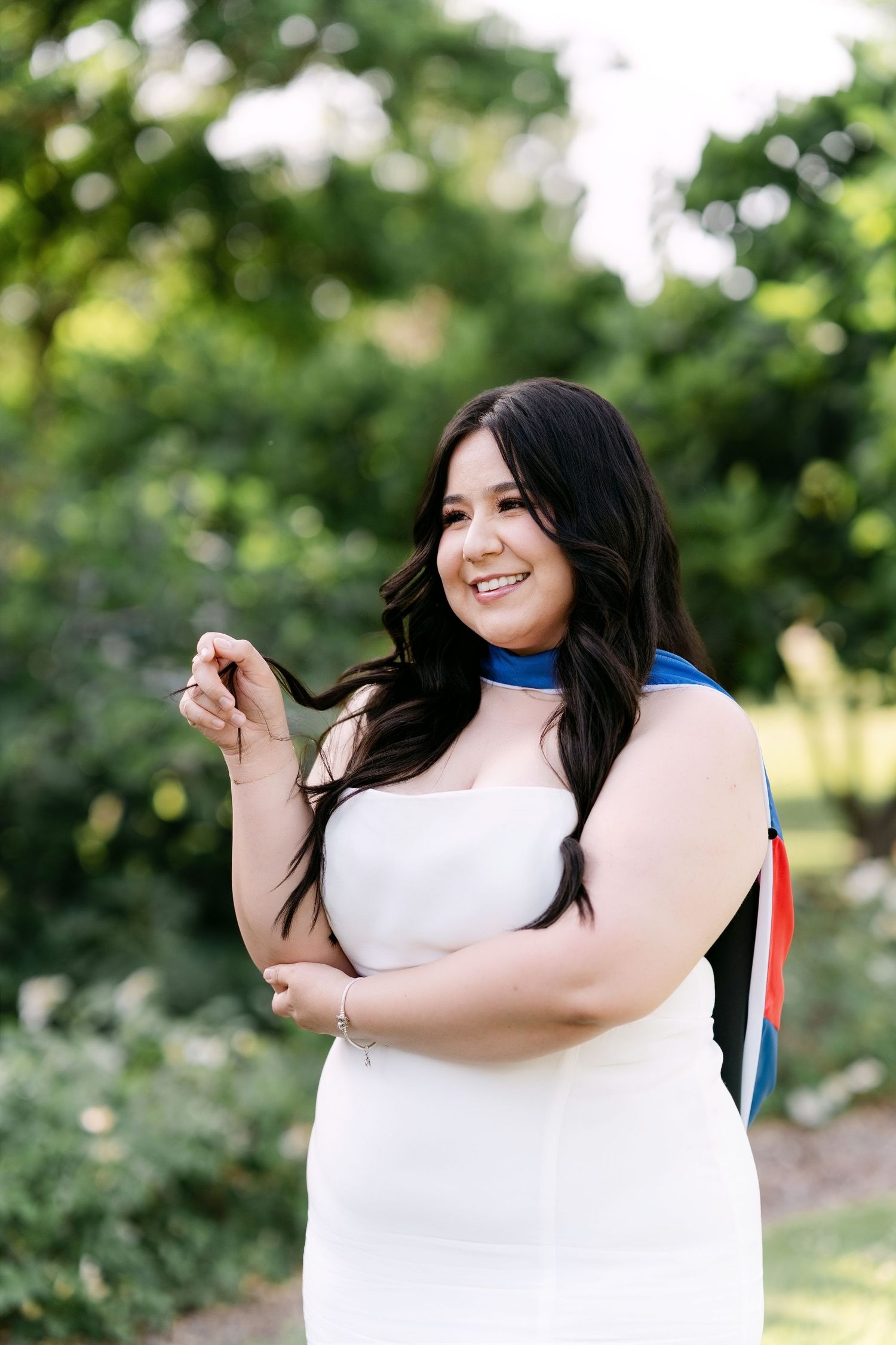 Smiling woman in white strapless dress with blue sash posing outdoors among green trees.