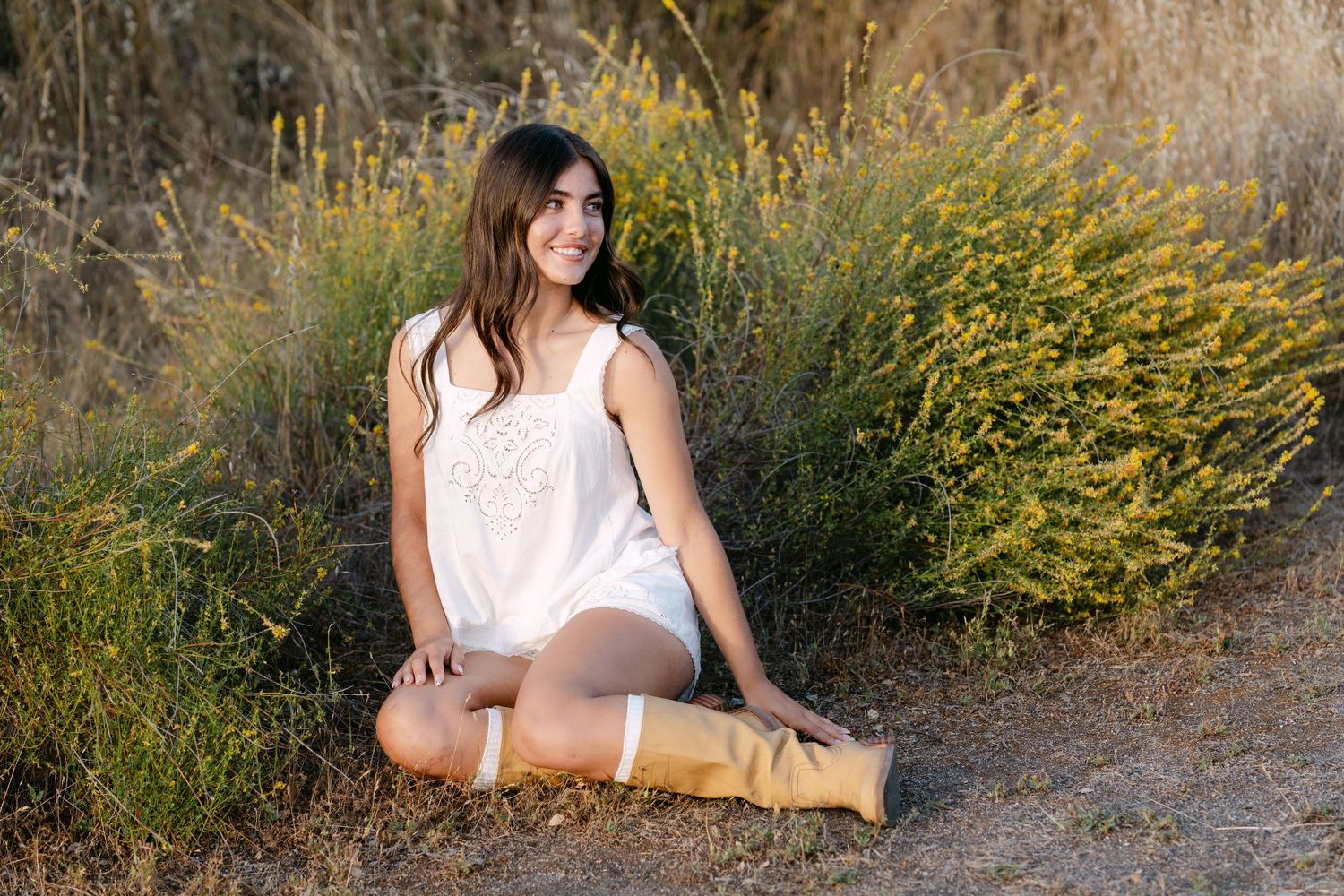 Young woman in white top and knee-high boots sitting outdoors on a dirt path surrounded by yellow wildflowers.