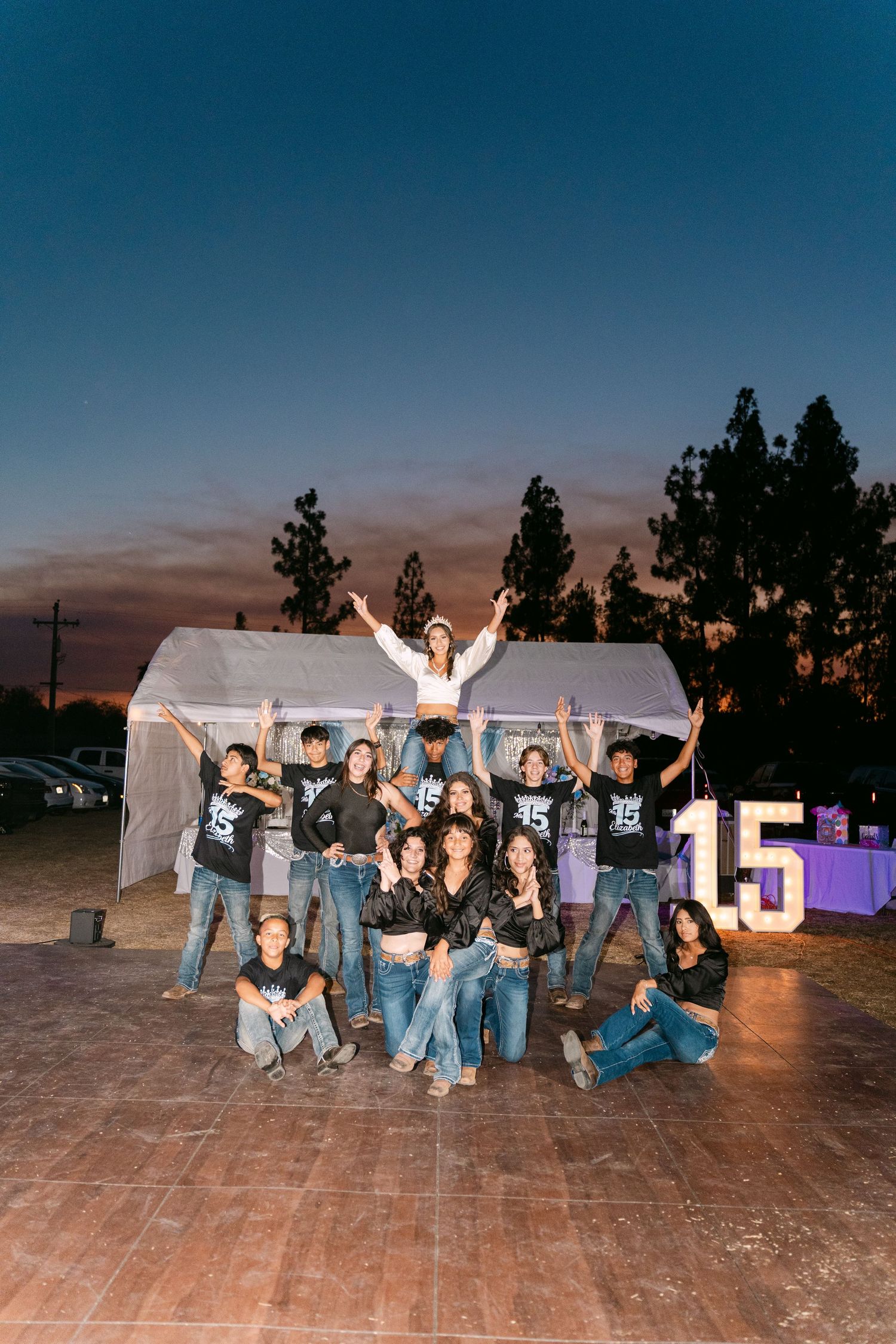 A celebratory group poses in front of a white tent with illuminated number 15 at dusk.