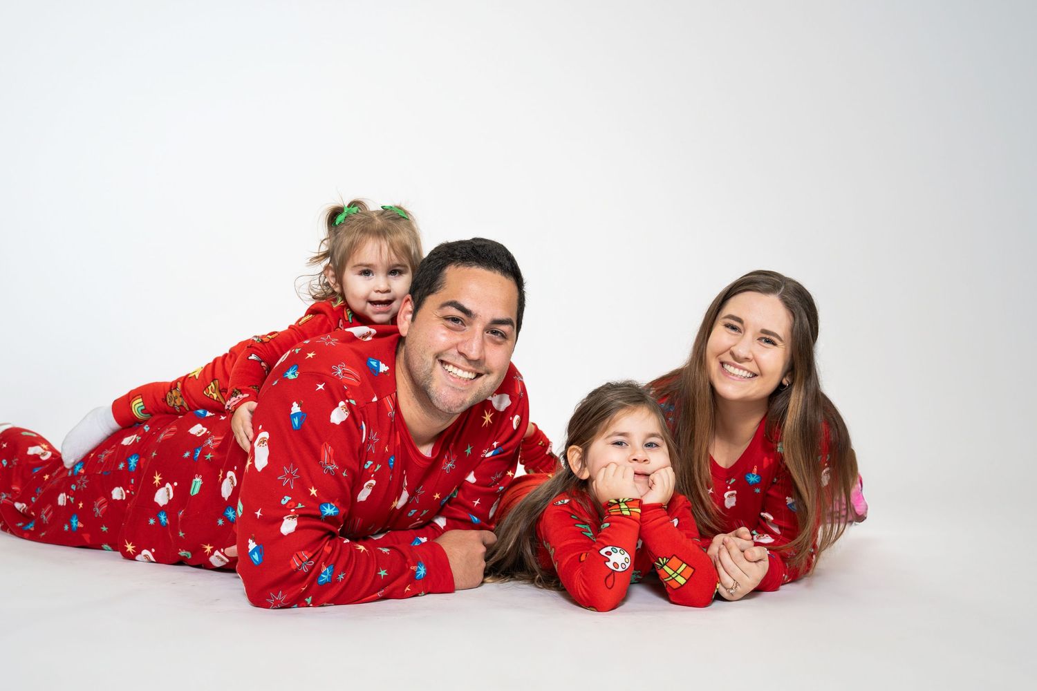 Family in matching red Christmas pajamas lying together on white floor.