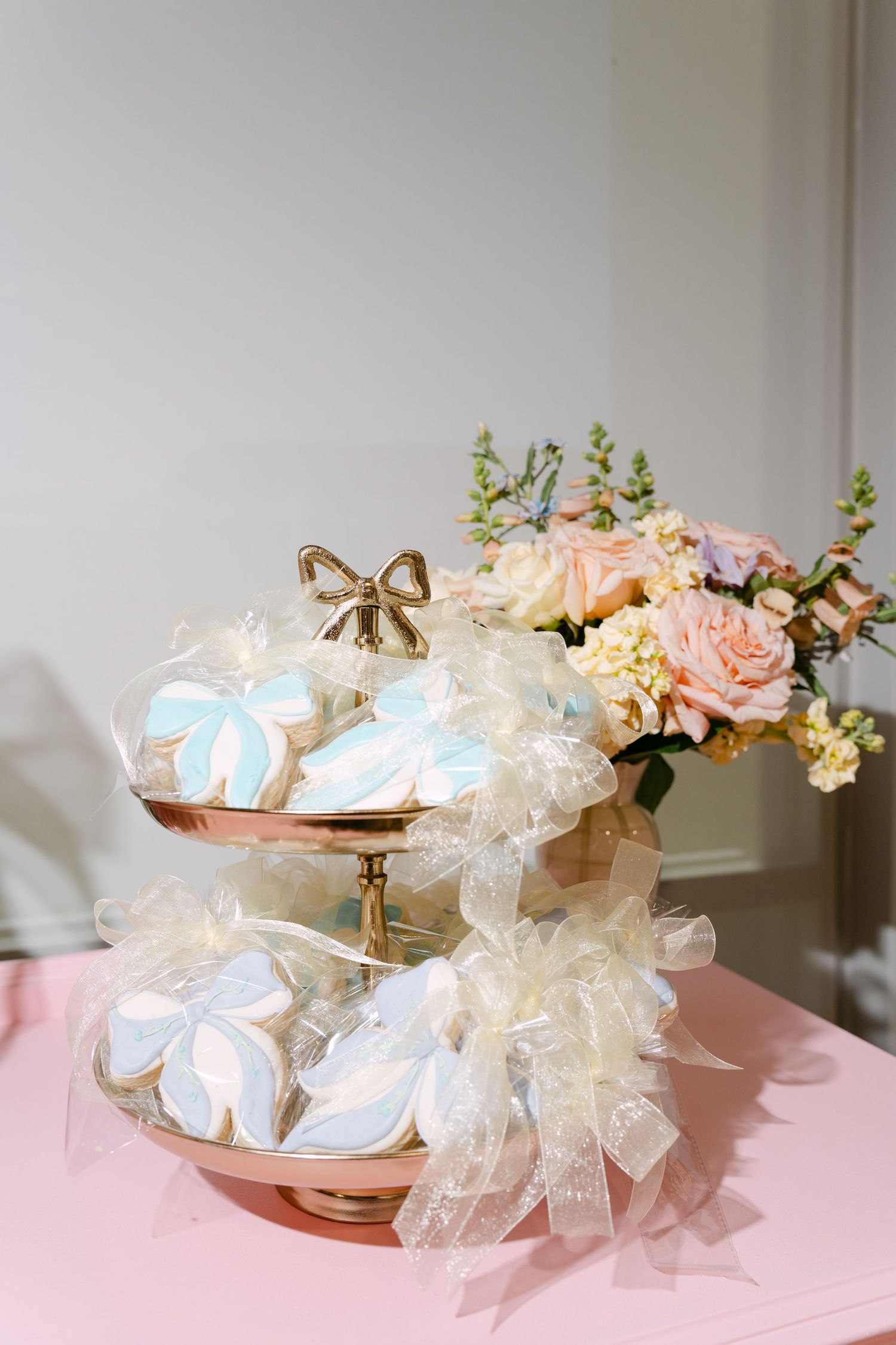 Elegant two-tier gold stand with blue bow cookies and cream ribbons beside a pastel floral arrangement on a pink table.