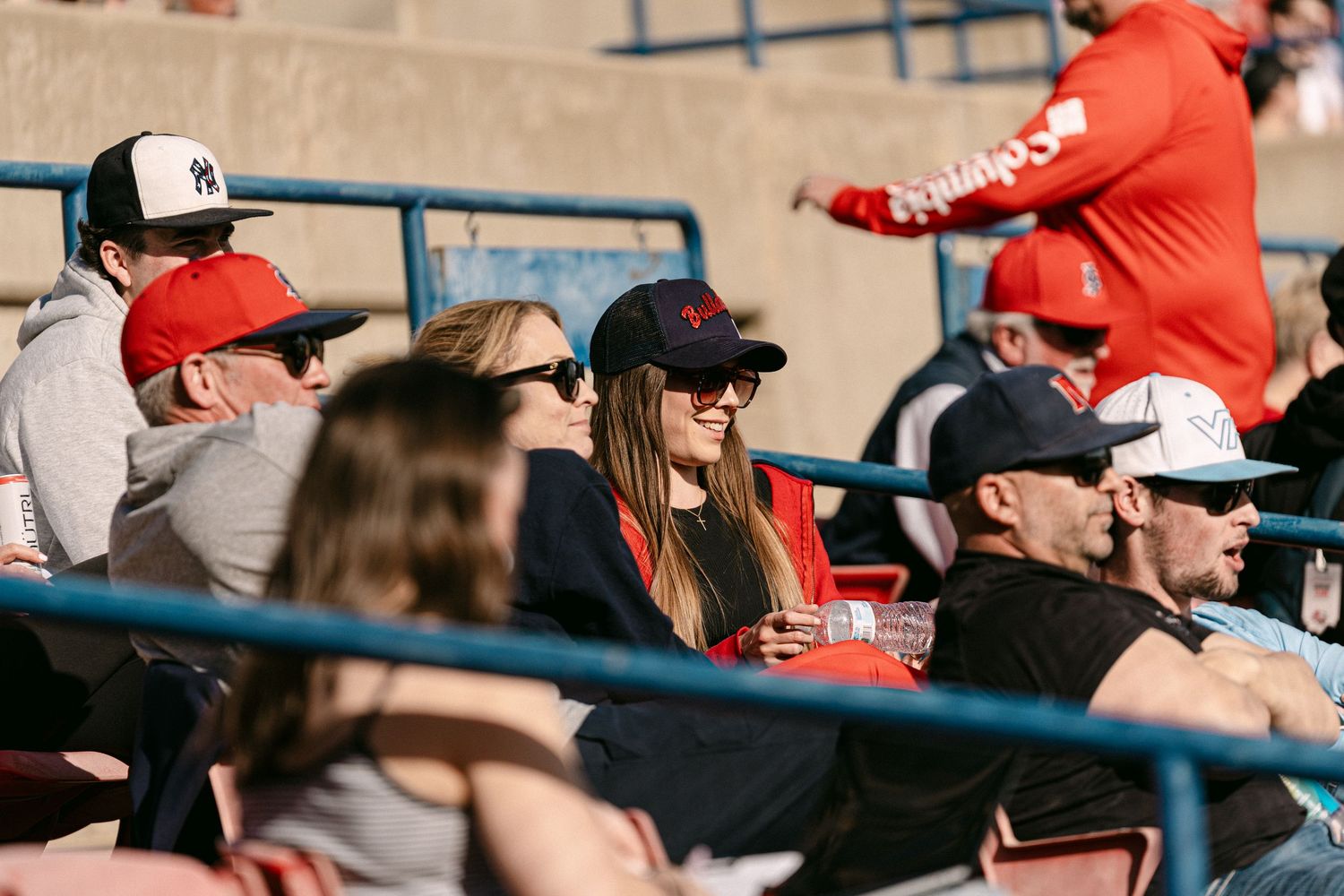 Baseball fans in red caps and sunglasses watch intently from stadium seats during a sunny afternoon game.