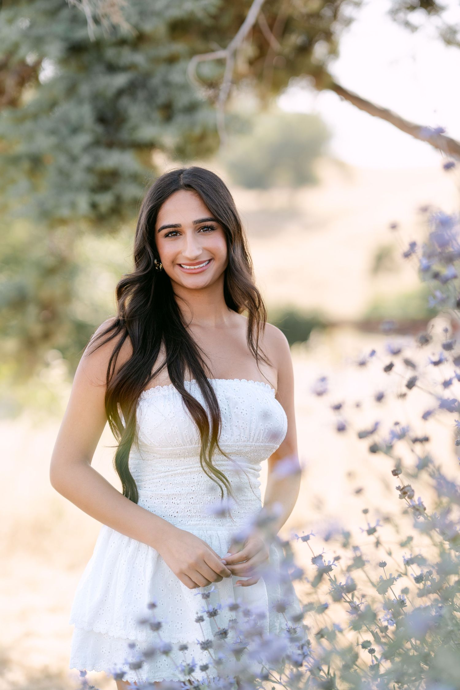 Young woman in white strapless dress smiling among lavender flowers in a sunlit outdoor field.