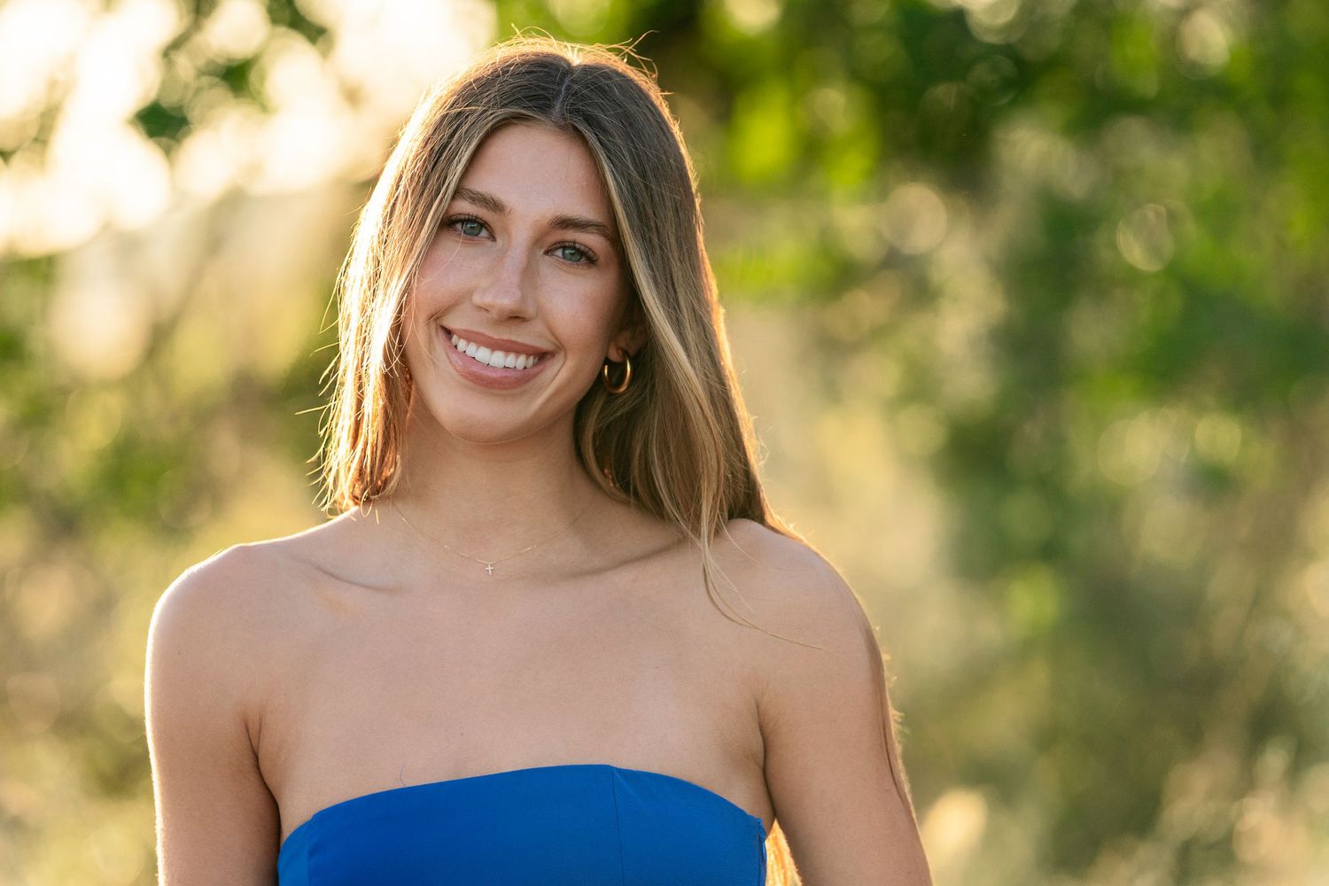High School Senior in blue strapless dress smiles during outdoor portrait session with bokeh background.