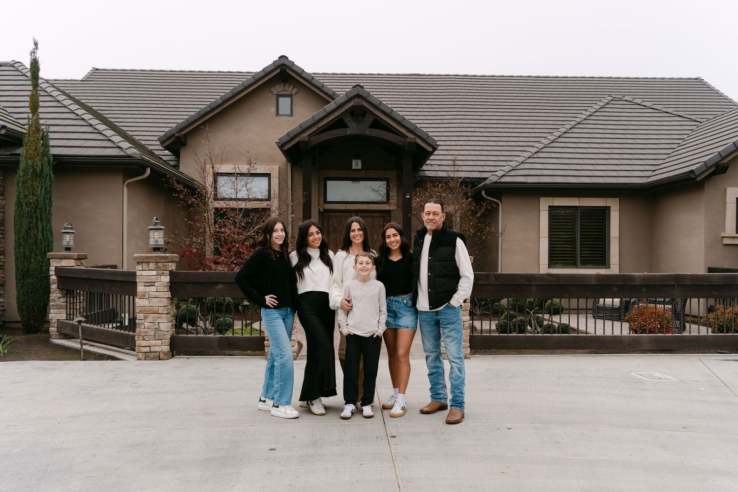 A family group poses together in front of their tan stucco house with stone accents.