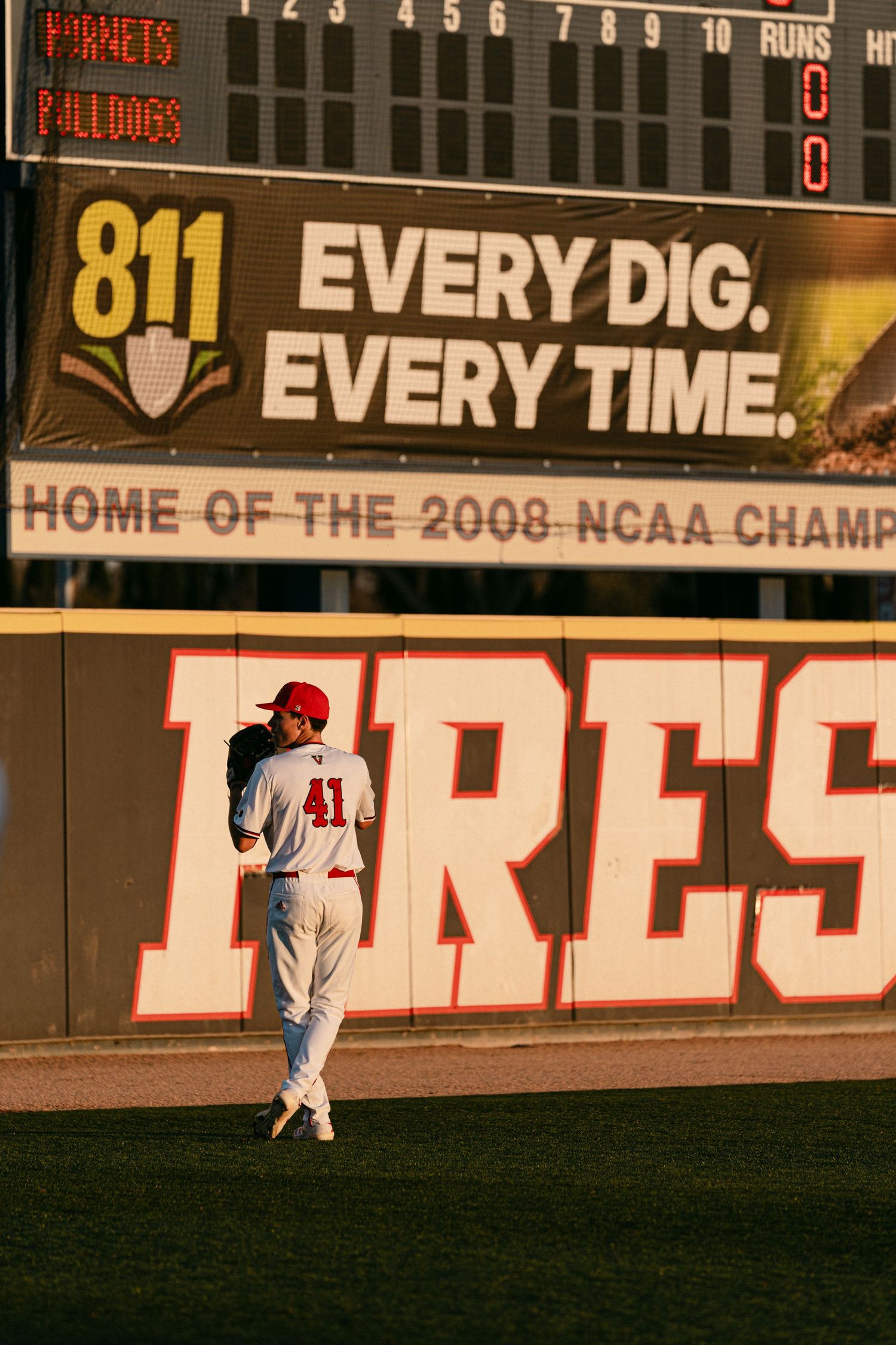 Baseball player stands near outfield wall with scoreboard and '811 EVERY DIG, EVERY TIME' banner at NCAA championship stadium.
