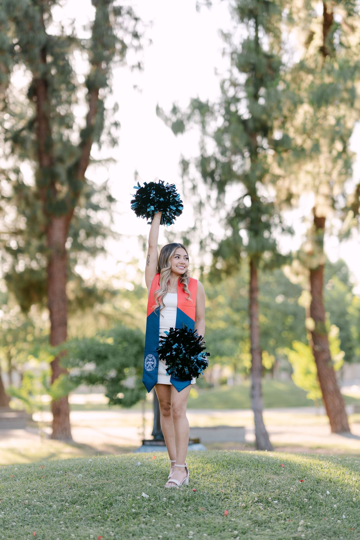 Cheerleader in red and blue uniform holds black pom-poms aloft outdoors, wearing graduation stole among tall trees.