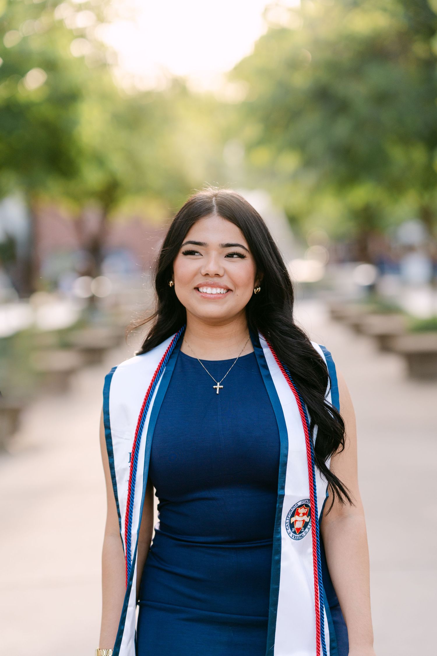 Smiling graduate in blue dress wearing white graduation stole with cross necklace outdoors on sunny campus.