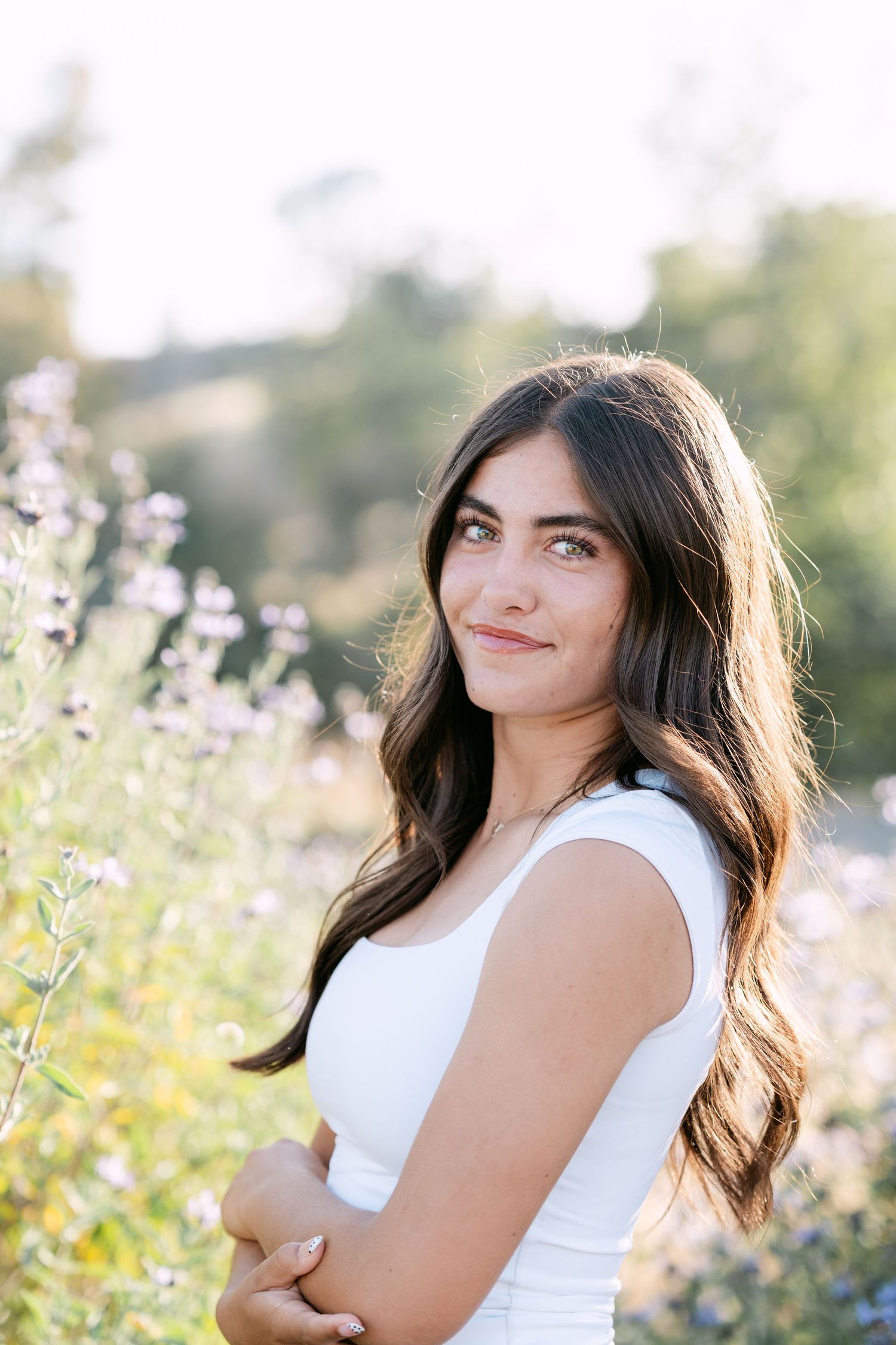 Young woman with long dark hair in white top smiling among purple wildflowers in sunny outdoor setting.
