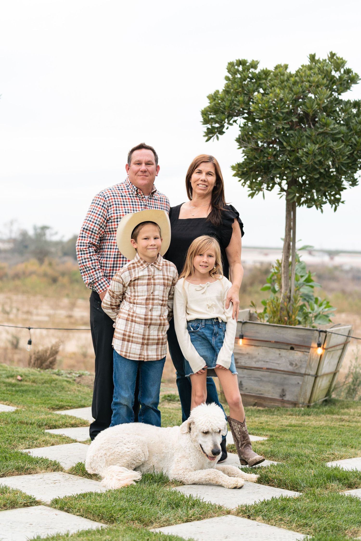 A family poses outdoors together with their white dog near wooden garden boxes on a grassy hillside.