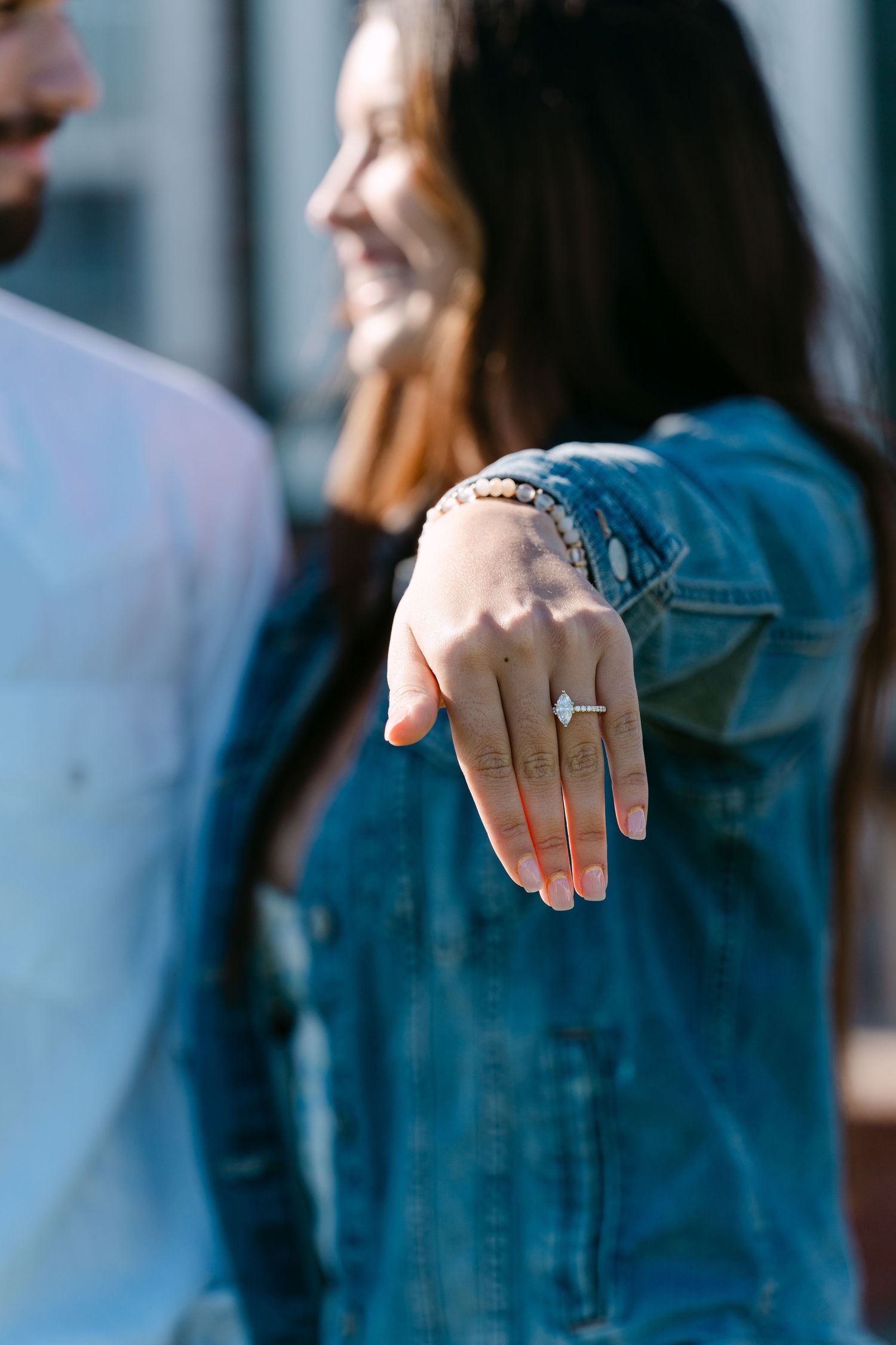 Someone wearing a denim jacket shows off their engagement ring with a joyful smile in the background.