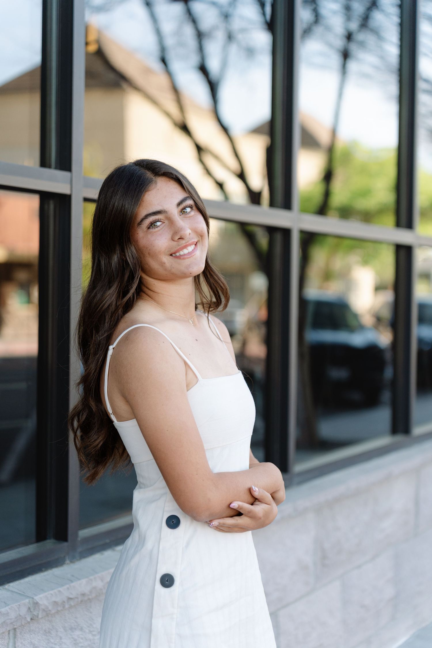 A person in a white sundress poses against a storefront window with black frames on a sunny day.