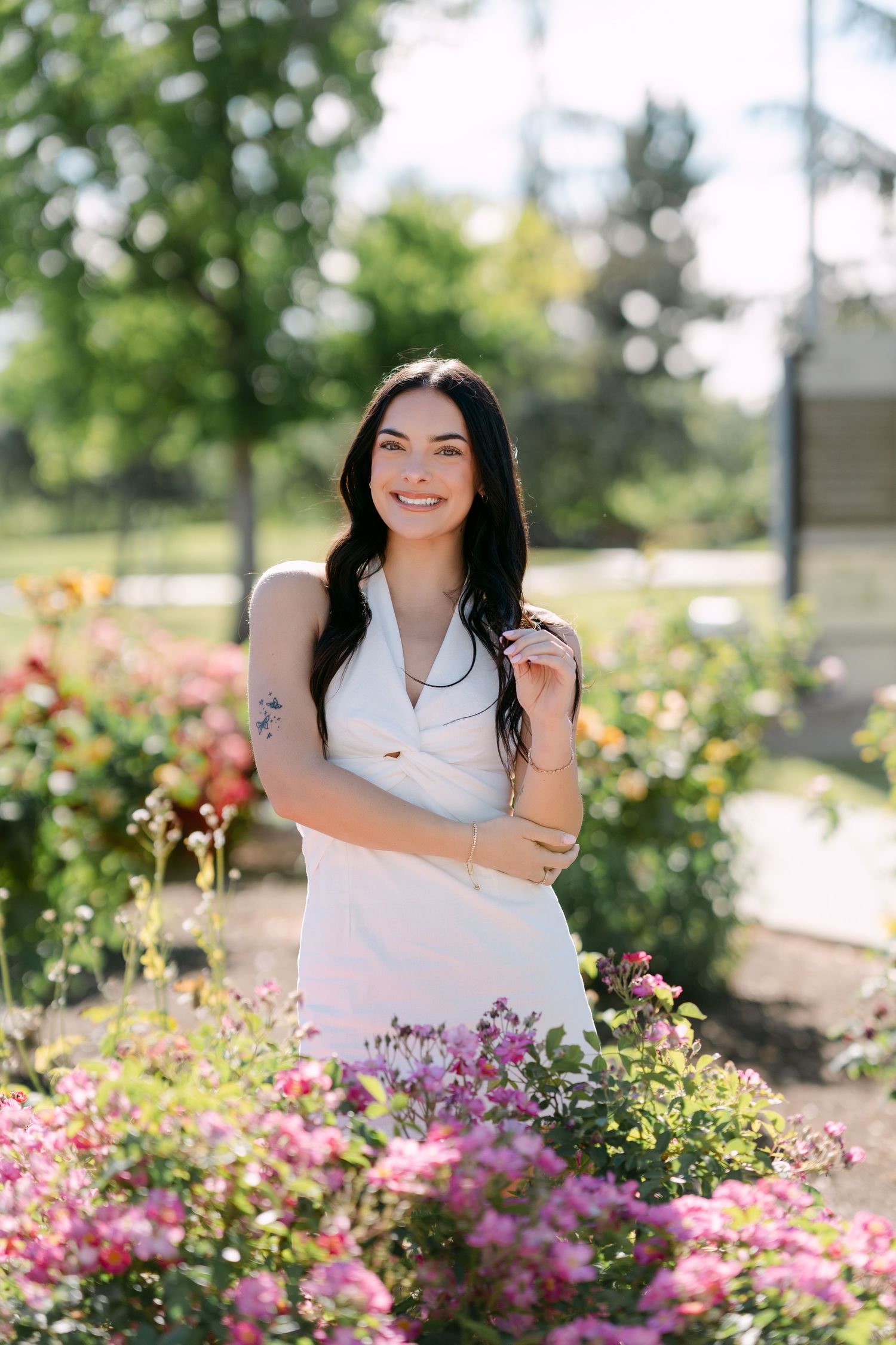 A smiling woman in a white dress stands among colorful blooming flowers in a sunny garden park.