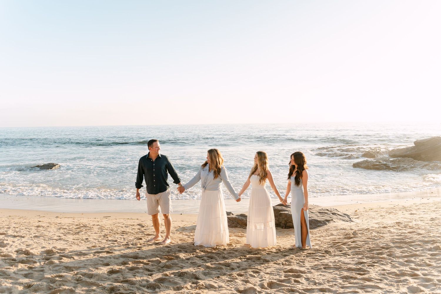Family of four holding hands on a sandy beach with ocean waves in the background at sunset.