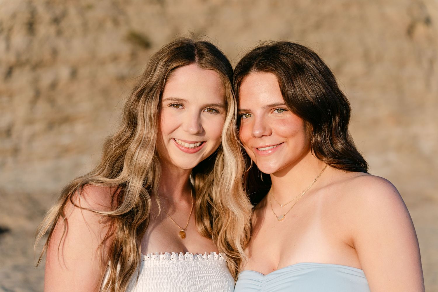 Two smiling young women with long hair pose together outdoors near a rocky backdrop in a warm golden light.